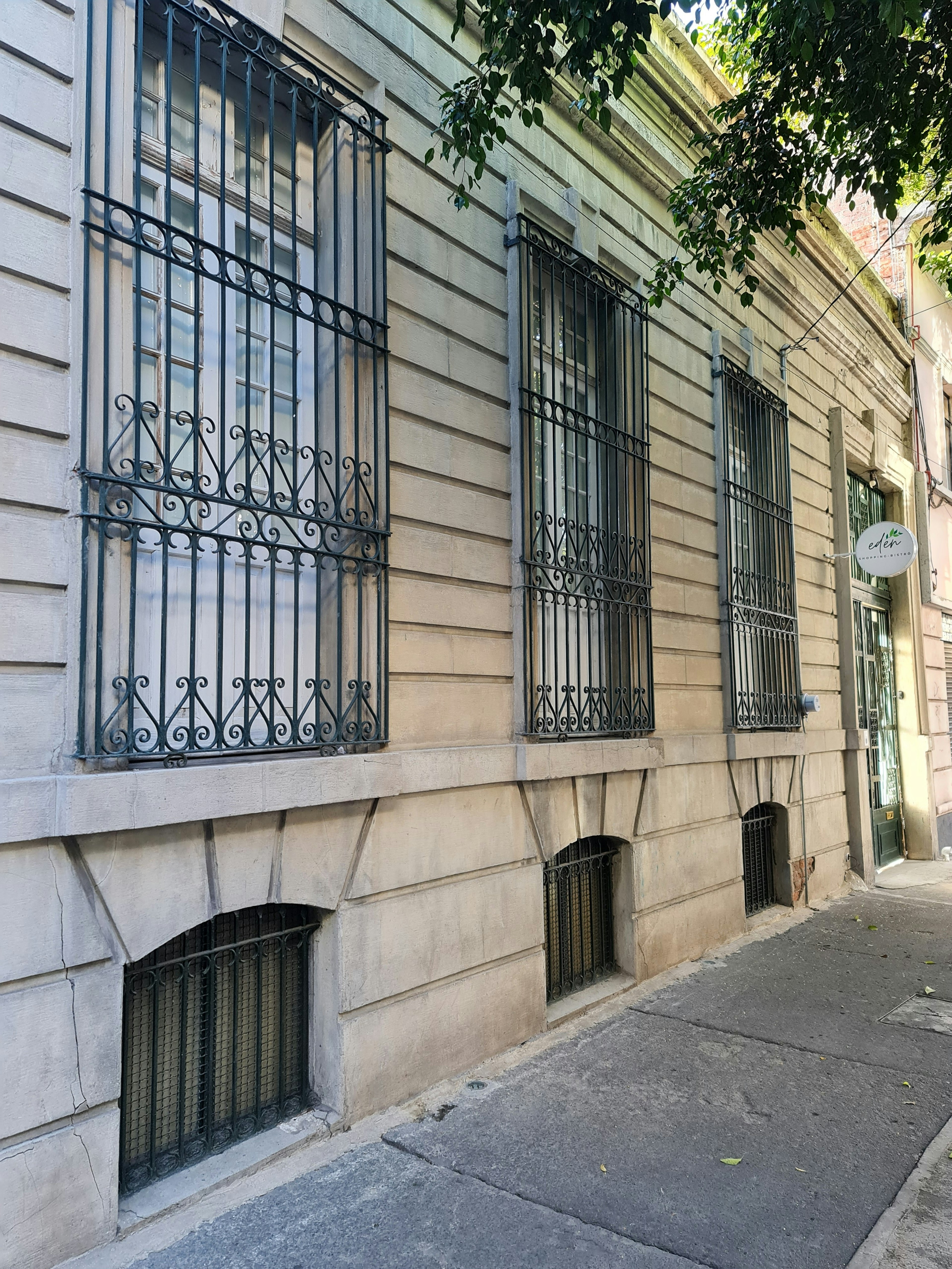 La imagen muestra la fachada de un edificio tradicional con elegantes ventanas protegidas por rejas de hierro forjado ornamentales. La construcción tiene un estilo arquitectónico clásico, con paredes de piedra lisa y detalles decorativos. Se observan también ventanas a nivel del suelo, probablemente de un sótano. El edificio parece estar bien mantenido y ubicado en una zona urbana, posiblemente residencial o comercial de alto nivel.