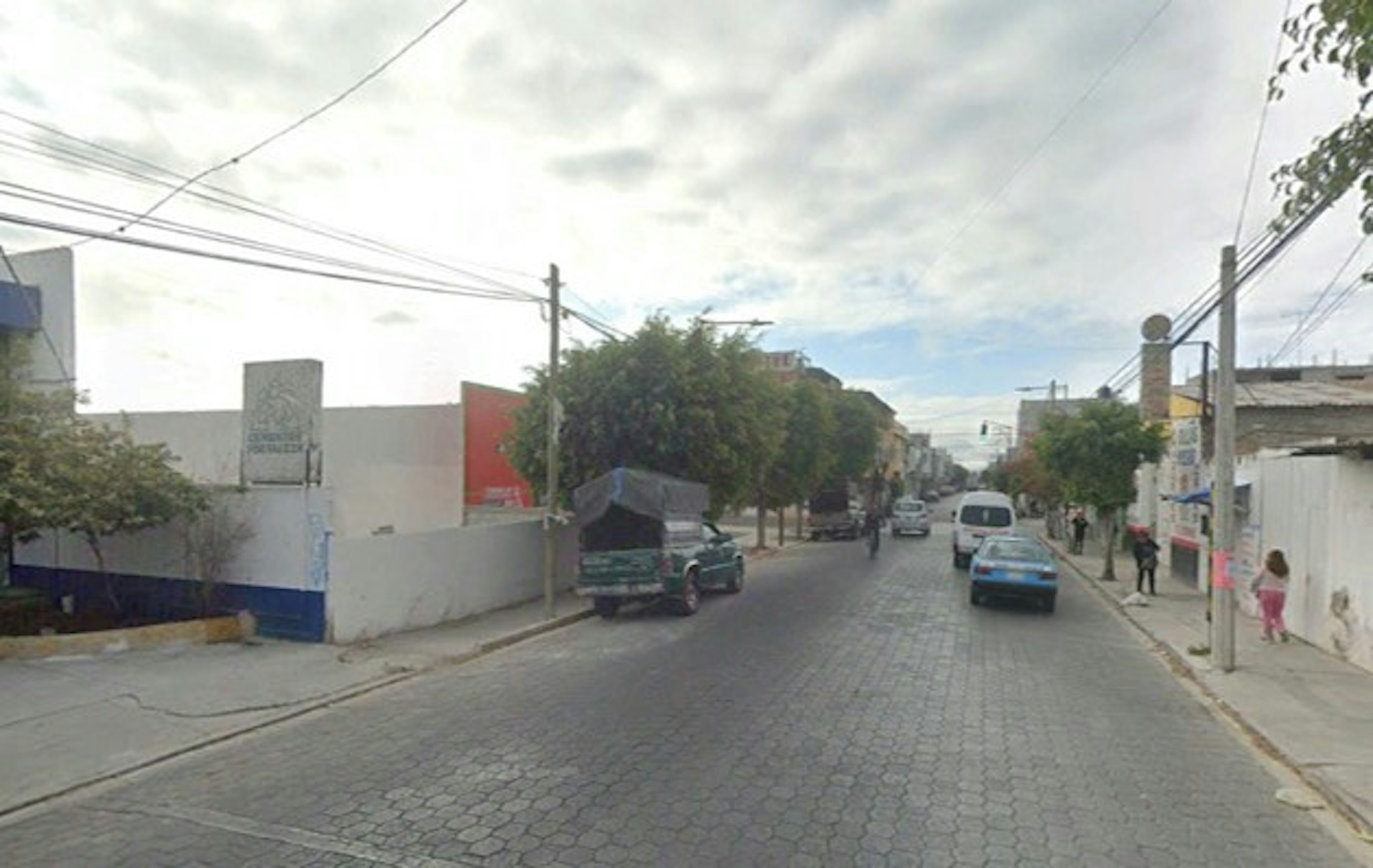 La imagen muestra una calle urbana con edificios bajos, árboles y aceras. El pavimento es de adoquines y hay algunos vehículos estacionados. Se ven cables eléctricos aéreos y postes. La calle parece tranquila, típica de una zona residencial o comercial de baja densidad. El estado general de las construcciones y la infraestructura sugiere una área en desarrollo o de clase media.