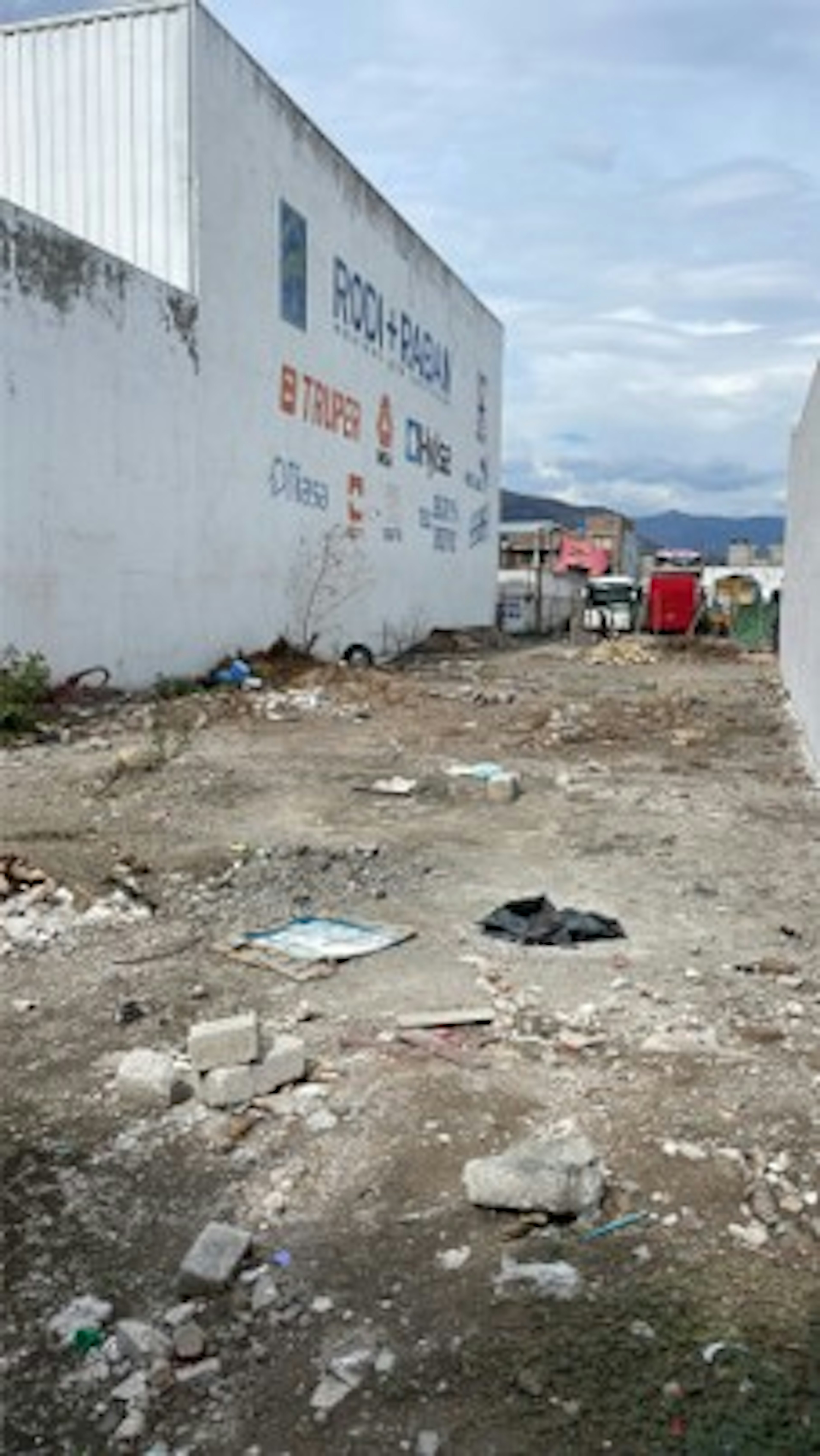 La imagen muestra un área urbana descuidada con un edificio blanco que tiene un letrero parcialmente visible. El terreno frente al edificio está lleno de escombros, basura y tierra suelta, indicando un posible sitio de construcción abandonado o un área en mal estado. El entorno sugiere una zona industrial o comercial de bajos recursos que podría necesitar rehabilitación o reurbanización.