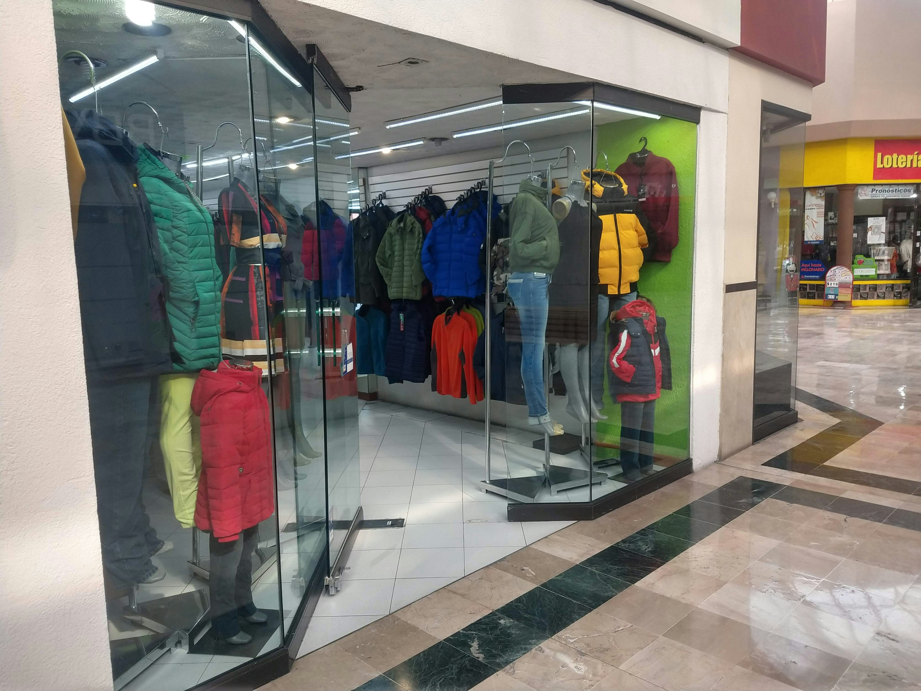 Vista de una tienda de ropa con maniquíes exhibiendo chaquetas y abrigos. Ubicada en un centro comercial, muestra un espacio comercial con vidrieras transparentes y un diseño interior moderno.