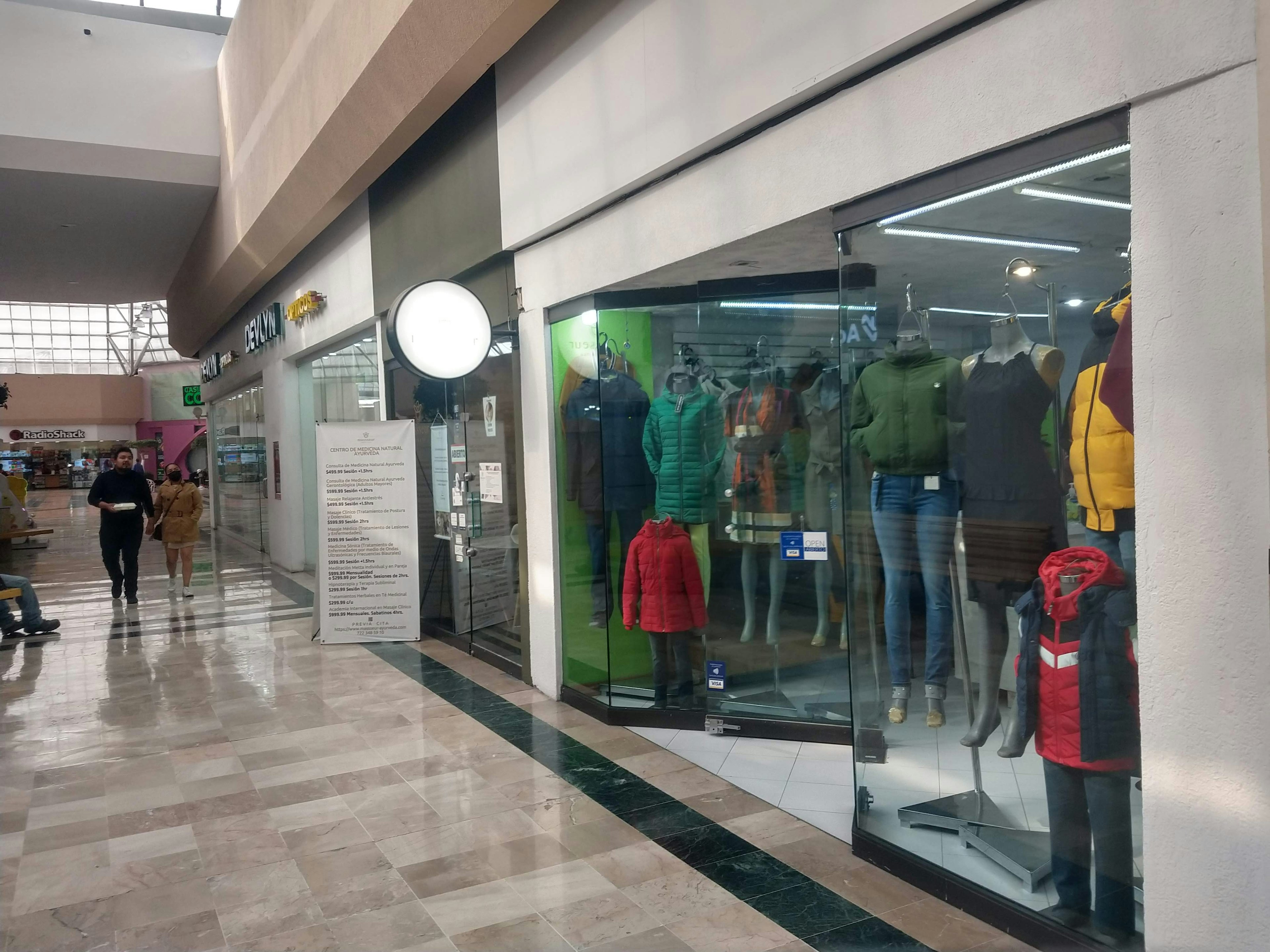 Imagen de un pasillo interior de un centro comercial con pisos de mármol pulido y tiendas a los lados. Se observa una tienda de ropa con maniquíes exhibiendo prendas y personas caminando.