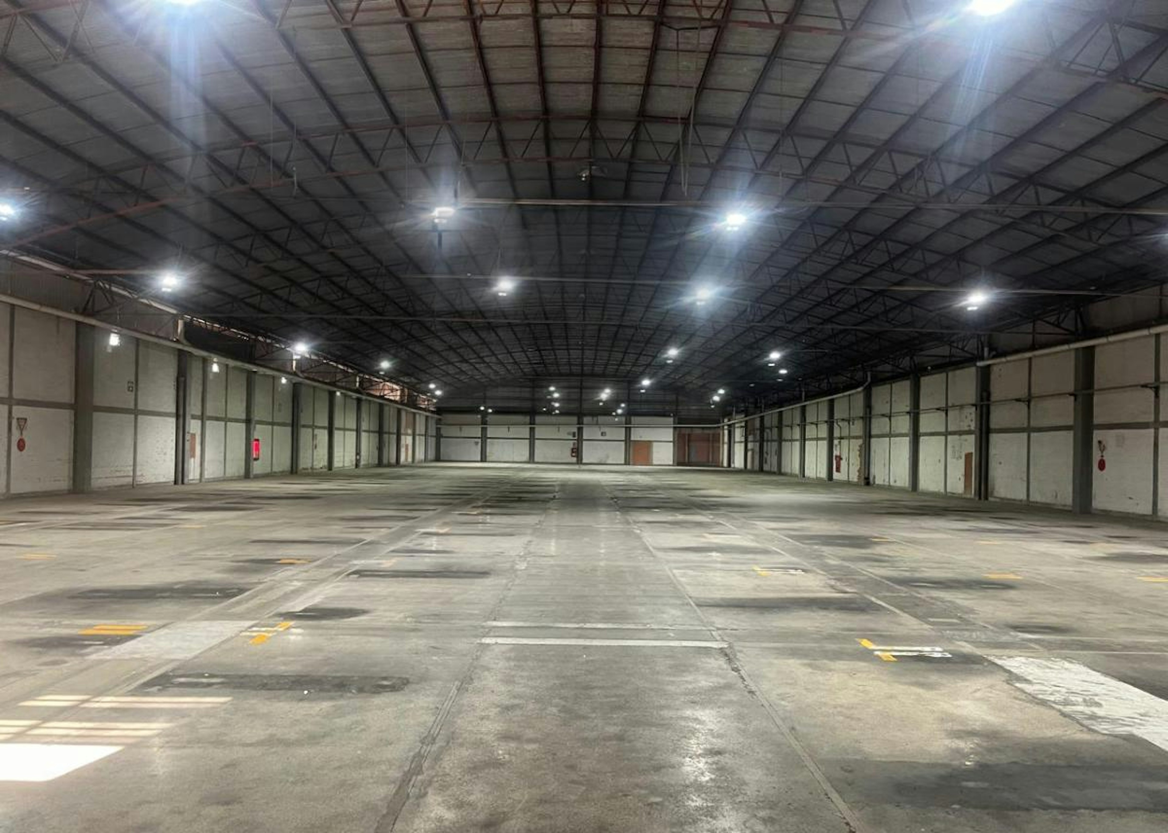 La imagen muestra un amplio almacén o nave industrial vacía con techo alto y estructura metálica. El espacio está bien iluminado con luces industriales. El suelo es de concreto con marcas amarillas. En las paredes laterales se ven puertas y paneles. Sobre el espacio se proyecta un gran logotipo transparente con un diseño que incluye un avión y formas geométricas, posiblemente el logo de la empresa propietaria del espacio.