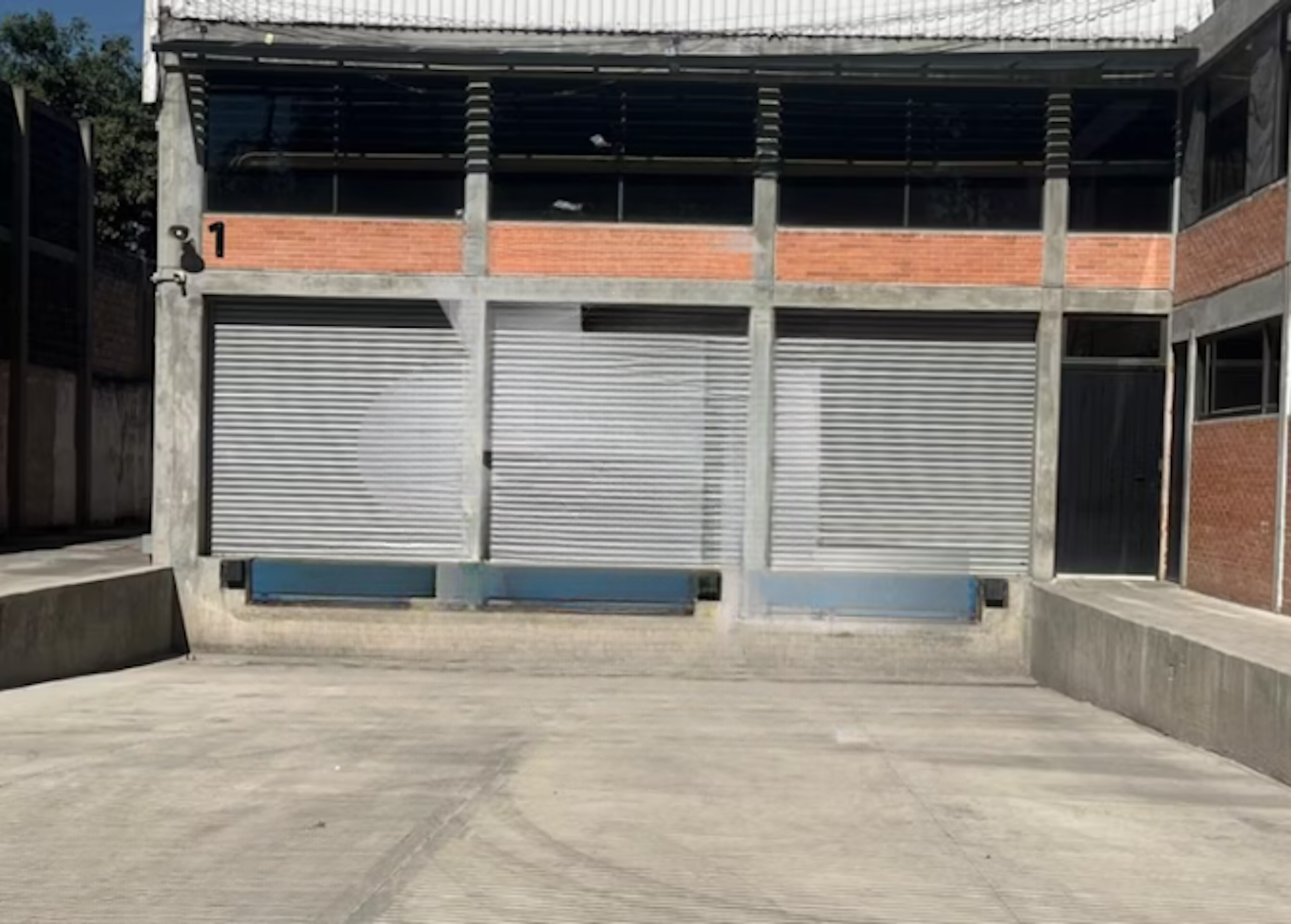 La imagen muestra un edificio comercial o industrial con una fachada de concreto y ladrillo. Tiene tres grandes puertas enrollables metálicas en la planta baja, numeradas con un 1 visible. La parte superior tiene ventanas oscuras y un techo de metal. El edificio parece estar en una zona industrial o de almacenes, con un patio de concreto en frente. La estructura combina elementos funcionales y estéticos típicos de propiedades comerciales.