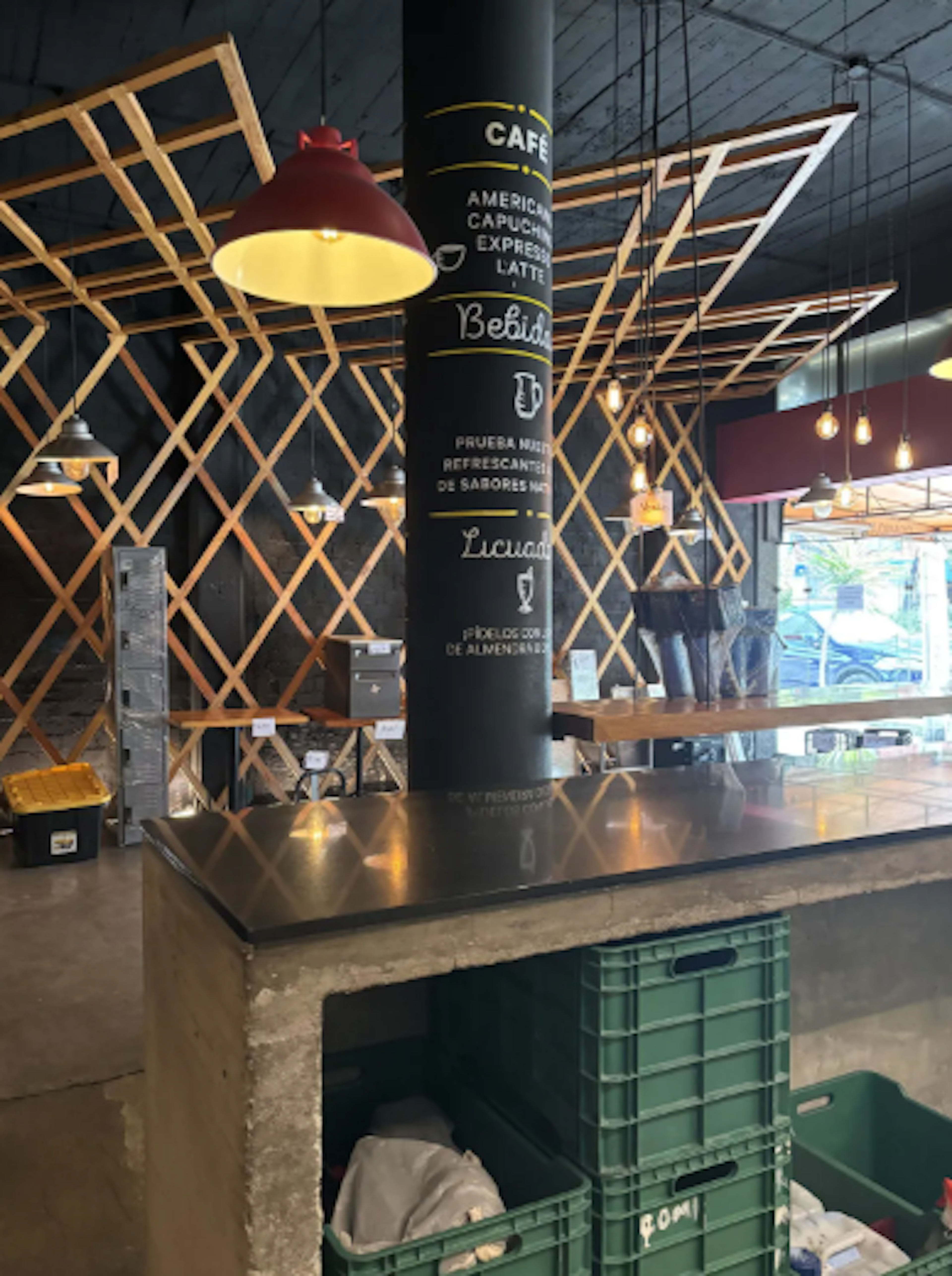 La imagen muestra el interior de un moderno café con un diseño industrial y acogedor. Destaca una estructura de madera en forma de celosía en el techo, lámparas colgantes y un pilar negro con el menú. El mostrador es de concreto con una superficie negra. Debajo hay cajas de plástico verde. El espacio combina elementos rústicos y contemporáneos, creando un ambiente atractivo para un local comercial.