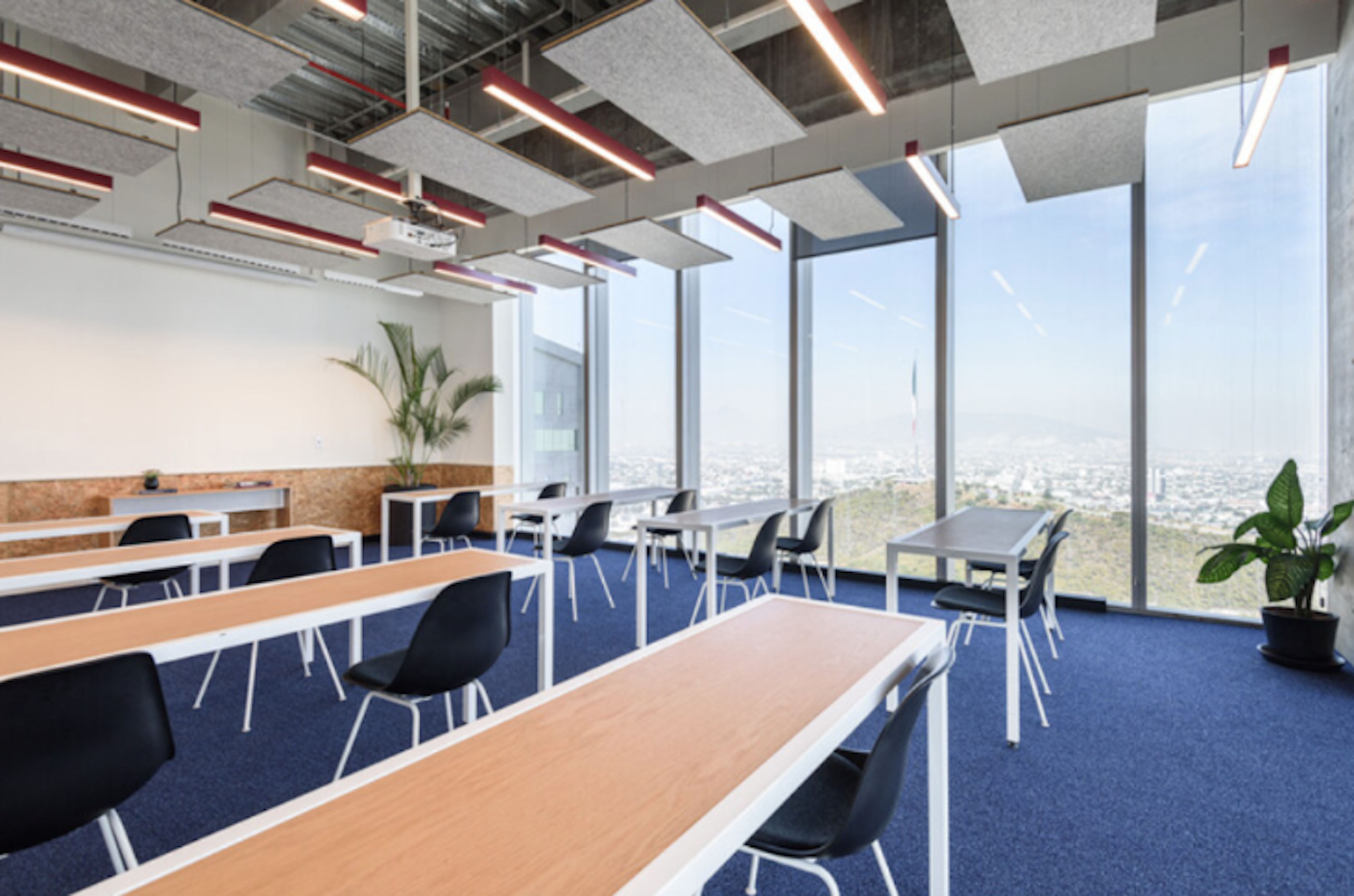 Este espacio de oficina moderno ofrece una vista panorámica de la ciudad a través de grandes ventanales. Cuenta con mesas de trabajo de madera, sillas negras ergonómicas y un suelo azul. El techo expuesto tiene iluminación LED roja. Las plantas y la decoración en corcho añaden un toque natural. El diseño abierto y luminoso crea un ambiente propicio para la productividad y la colaboración.