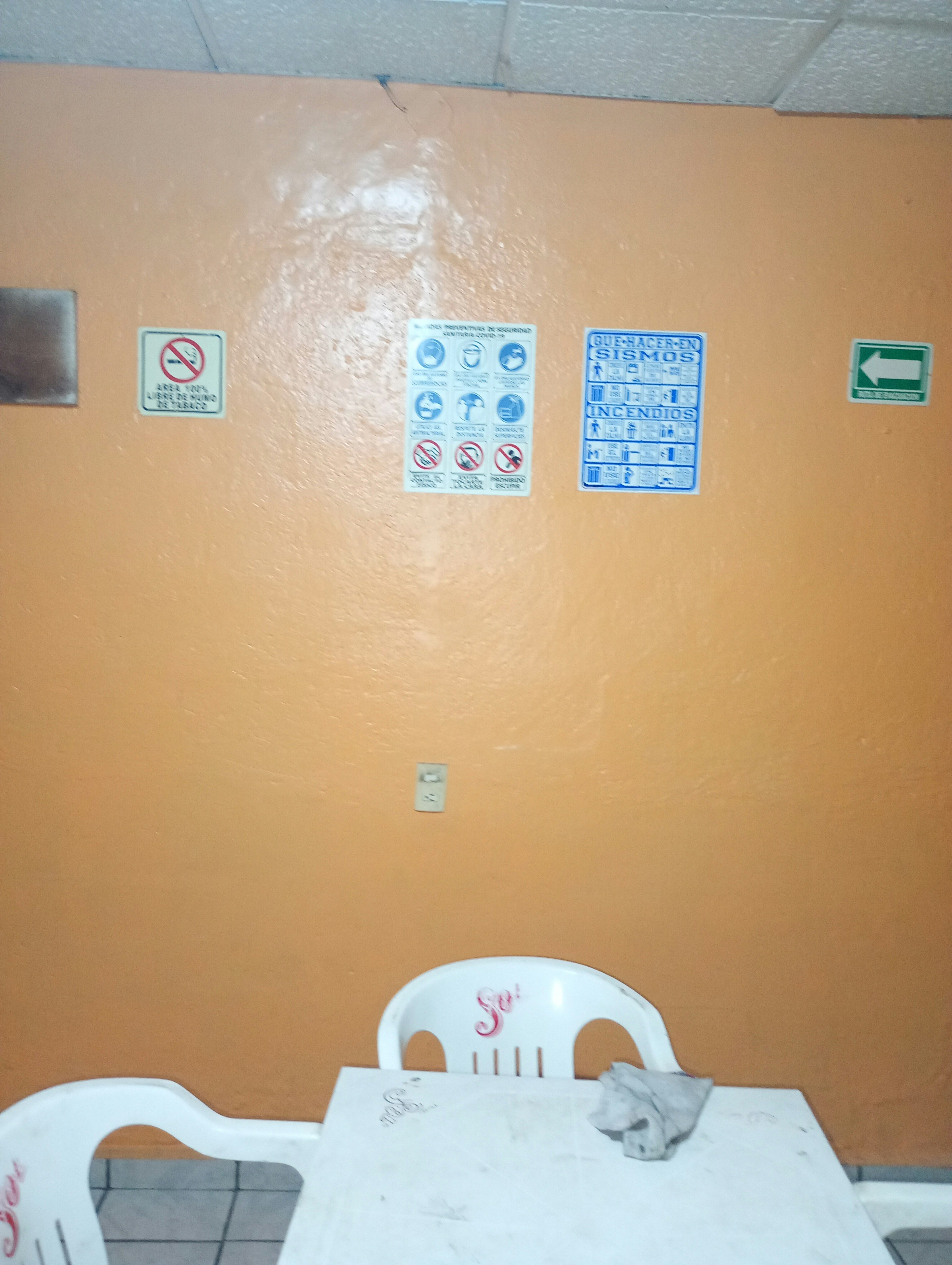 La imagen muestra una pared de color naranja con varios carteles de seguridad, incluyendo instrucciones para sismos e incendios. Hay un tomacorriente en la pared y en la parte inferior se ve una mesa blanca con sillas de plástico. El ambiente parece ser un espacio común o sala de espera en un edificio, posiblemente de oficinas o institución pública.