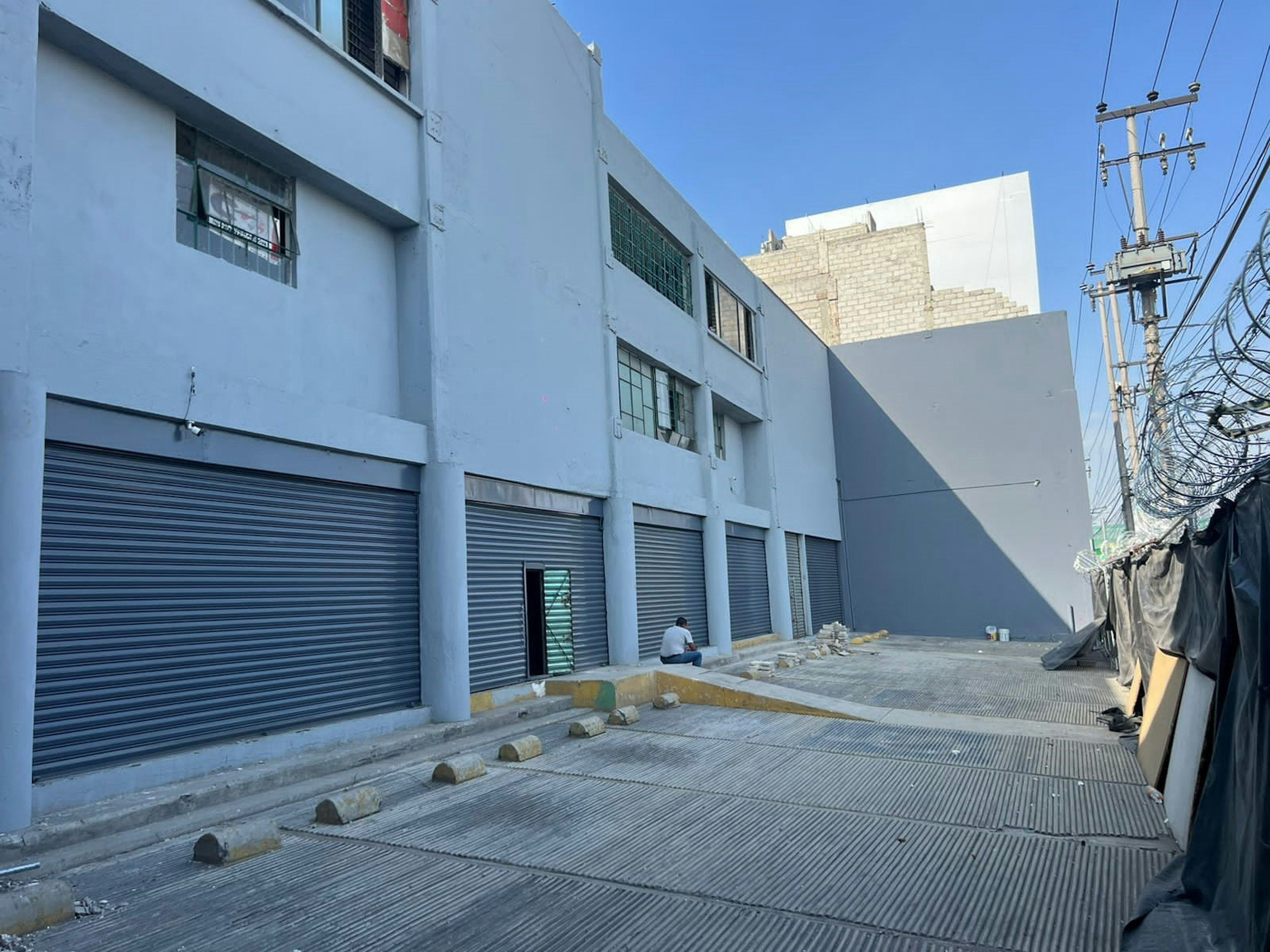 La imagen muestra un edificio industrial o comercial de dos pisos pintado de azul claro. Tiene varias puertas enrollables de metal cerradas en la planta baja, sugiriendo espacios de almacenamiento o tiendas. El edificio parece estar en una zona urbana, con cables eléctricos visibles y una calle pavimentada al frente. Se observa un área en construcción o remodelación, con algunos materiales y escombros en el suelo.