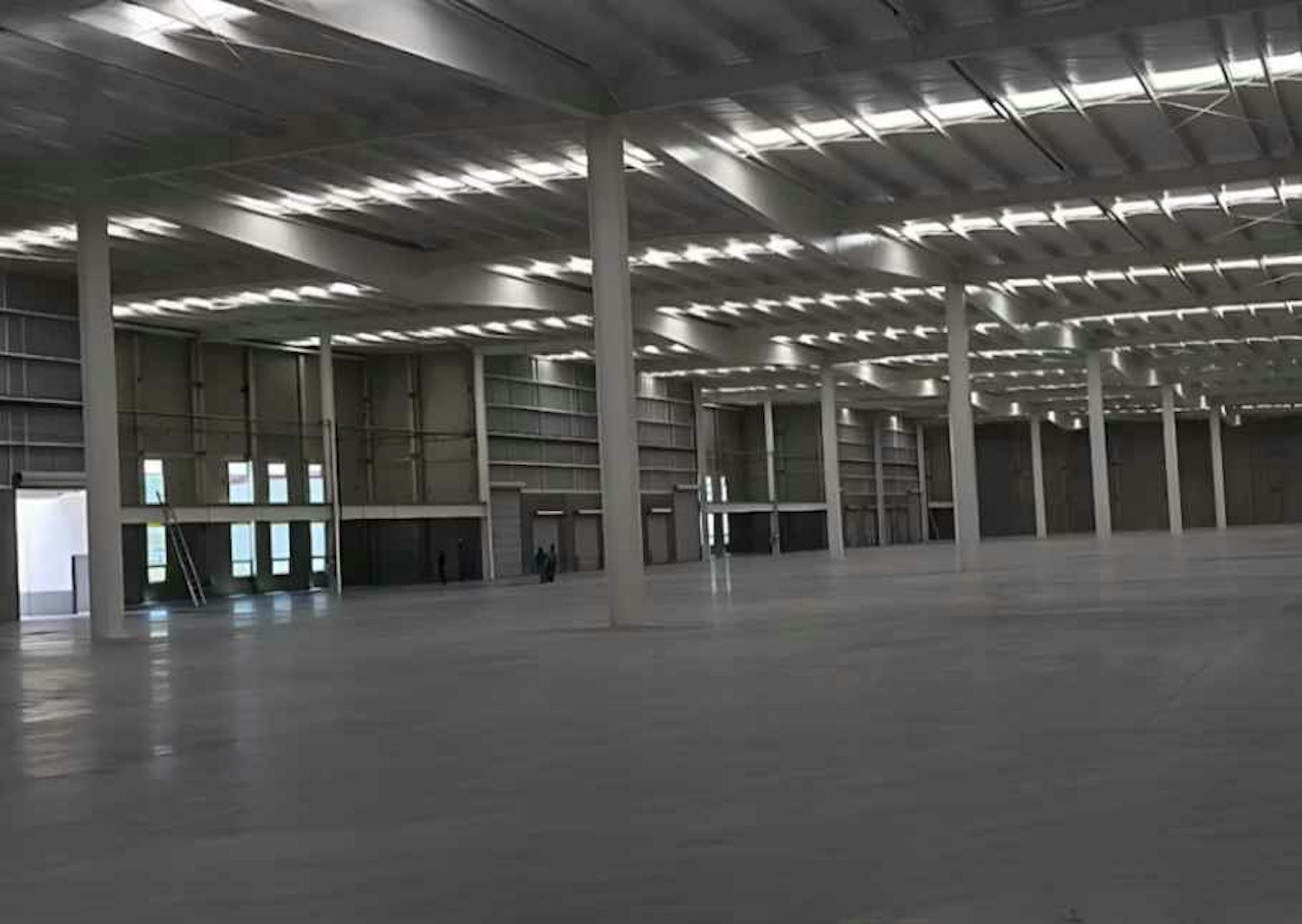 Esta imagen muestra un amplio espacio industrial o de almacenamiento. El interior cuenta con un techo alto sostenido por columnas blancas. El suelo es de concreto pulido y hay múltiples filas de iluminación fluorescente en el techo. Al fondo se pueden ver grandes puertas o ventanales. El espacio está vacío, lo que sugiere que está listo para ser utilizado o alquilado para fines comerciales o logísticos.