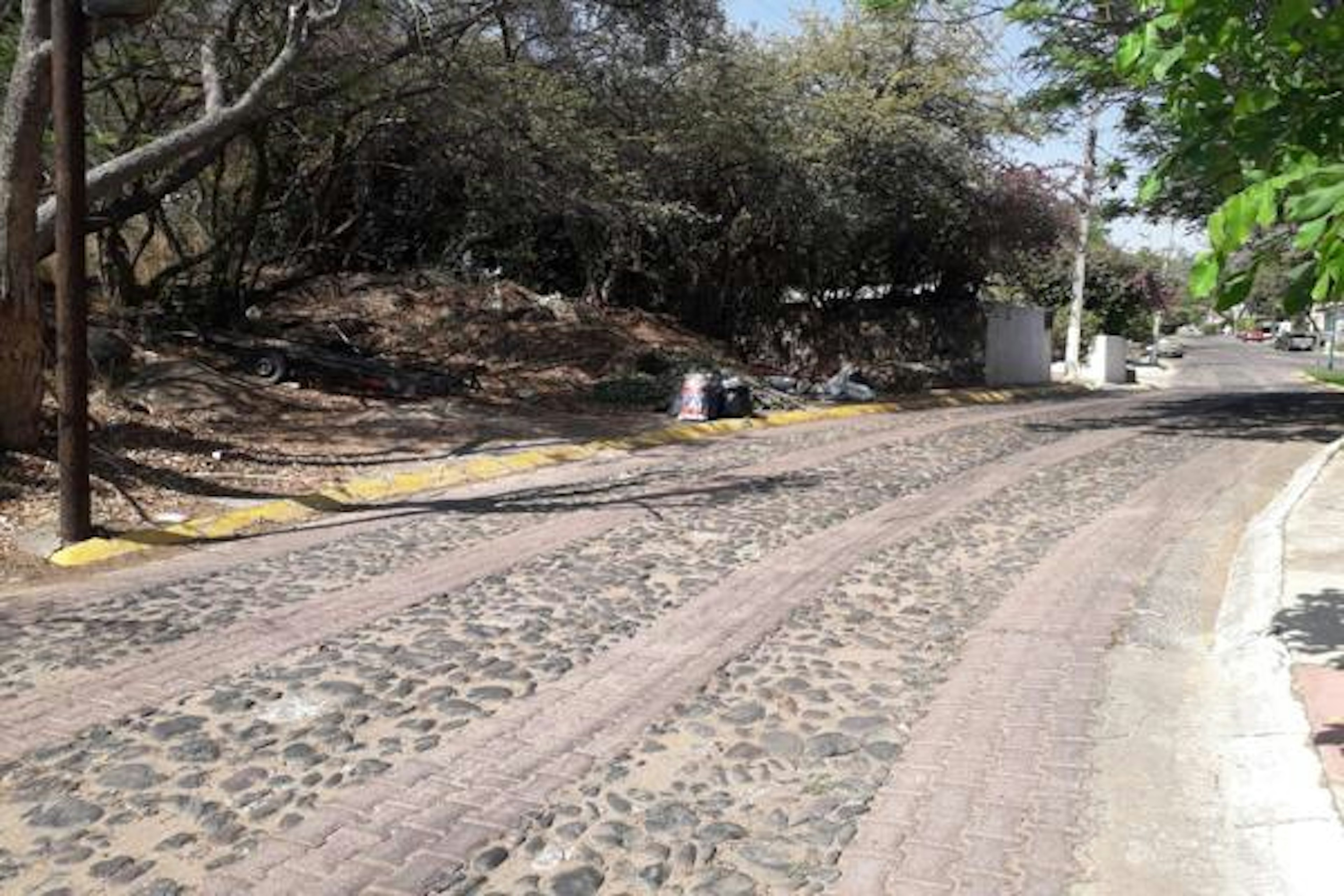 Imagen de una calle adoquinada con aceras y vegetación a los lados, posiblemente en una zona residencial. Se observa un terreno con árboles y arbustos al fondo. Ideal para destacar la tranquilidad y naturaleza del entorno.