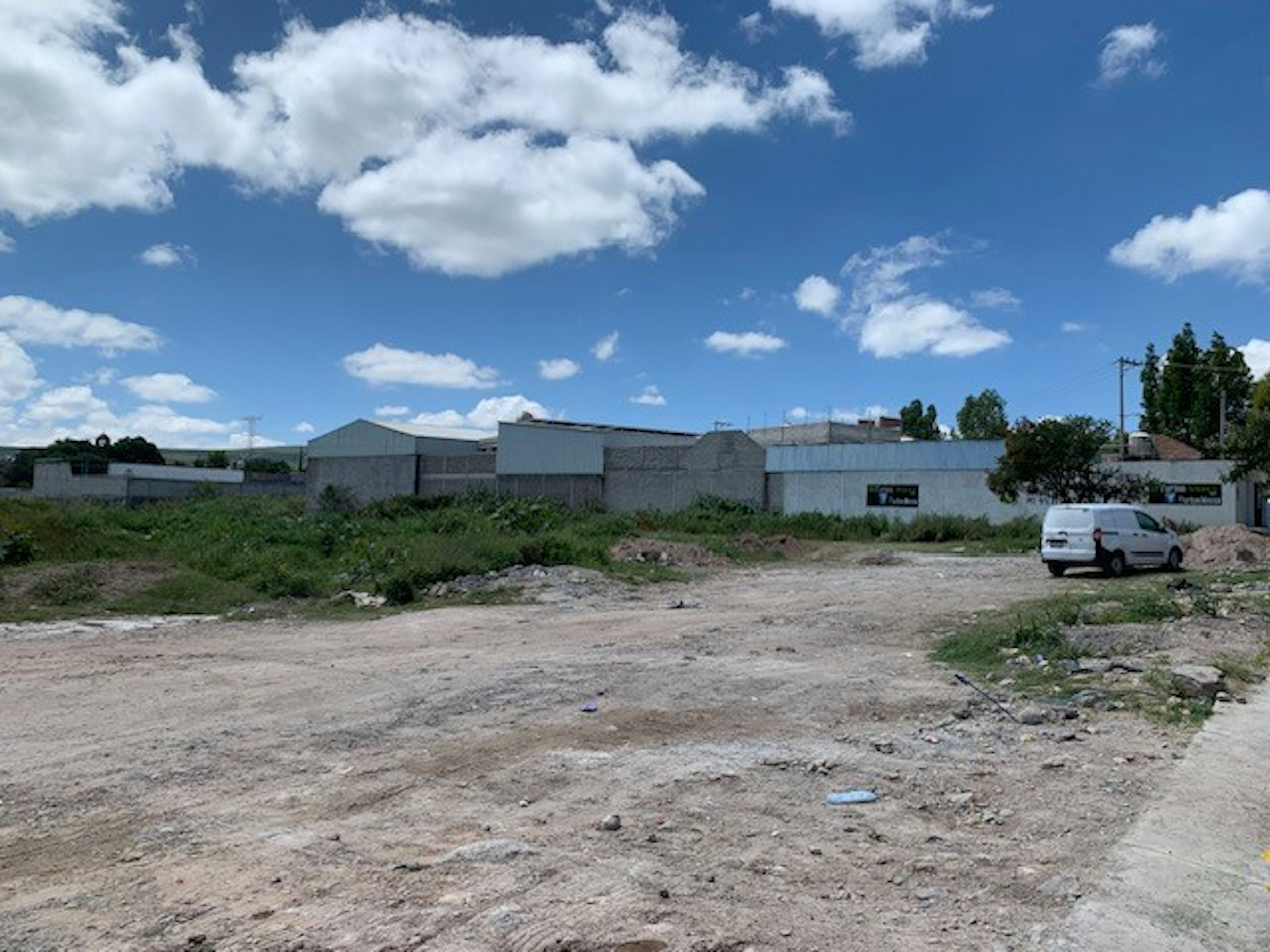 La imagen muestra un terreno baldío en un área suburbana o semi-rural. El cielo está despejado con algunas nubes blancas. En el fondo se ven edificaciones de baja altura con muros blancos, posiblemente almacenes o naves industriales. Hay vegetación dispersa y un vehículo utilitario blanco estacionado. El terreno parece estar sin desarrollar, con tierra suelta y algunos escombros, sugiriendo una potencial oportunidad de desarrollo inmobiliario.