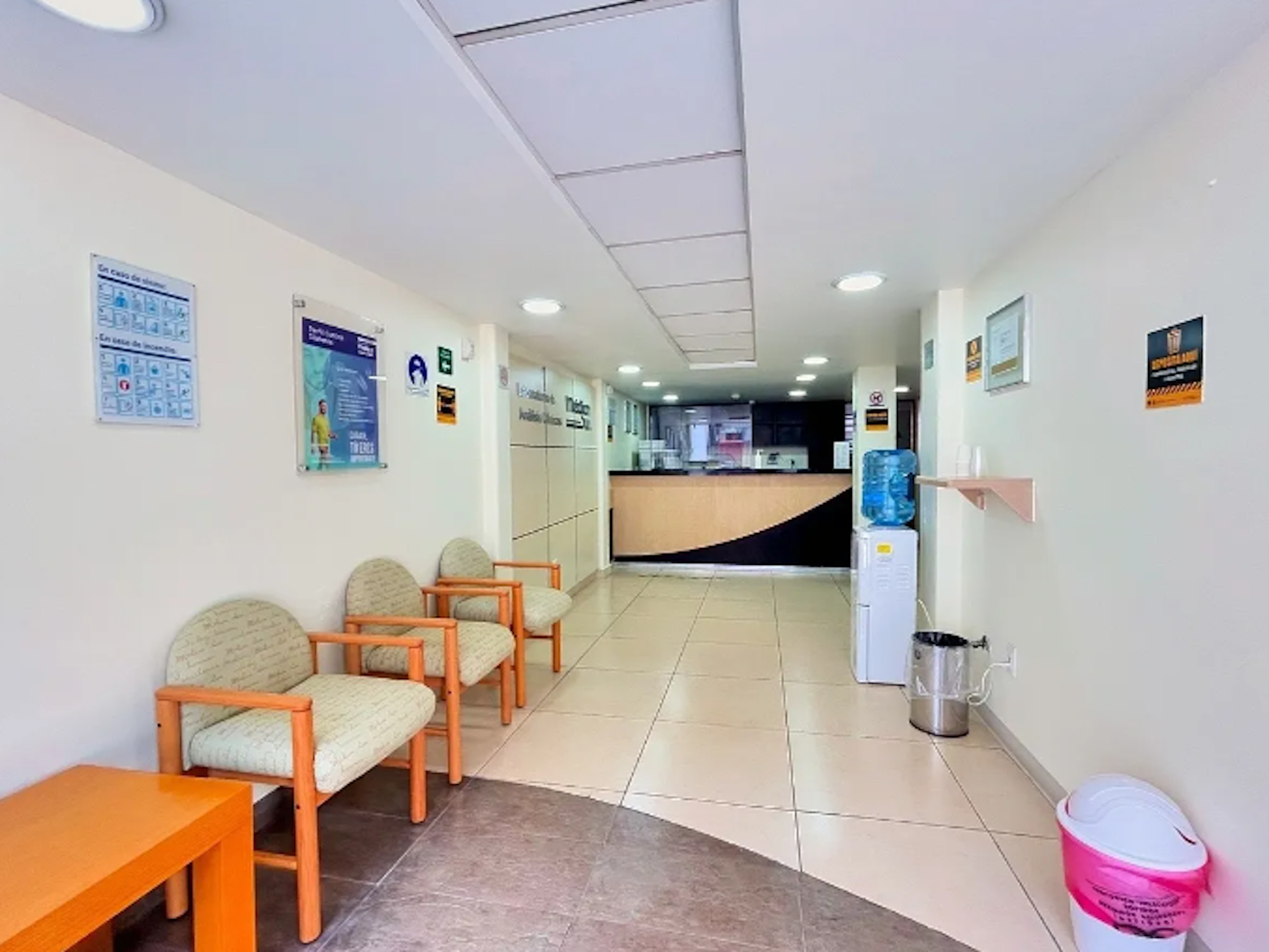 La imagen muestra el interior de una clínica médica o consultorio. Se observa una sala de espera con sillas acolchadas de madera, un mostrador de recepción al fondo, y paredes blancas con carteles informativos. El espacio está bien iluminado con luces empotradas en el techo y cuenta con un dispensador de agua y un bote de basura. El suelo es de baldosas claras y hay un ambiente limpio y ordenado, típico de un establecimiento de salud.