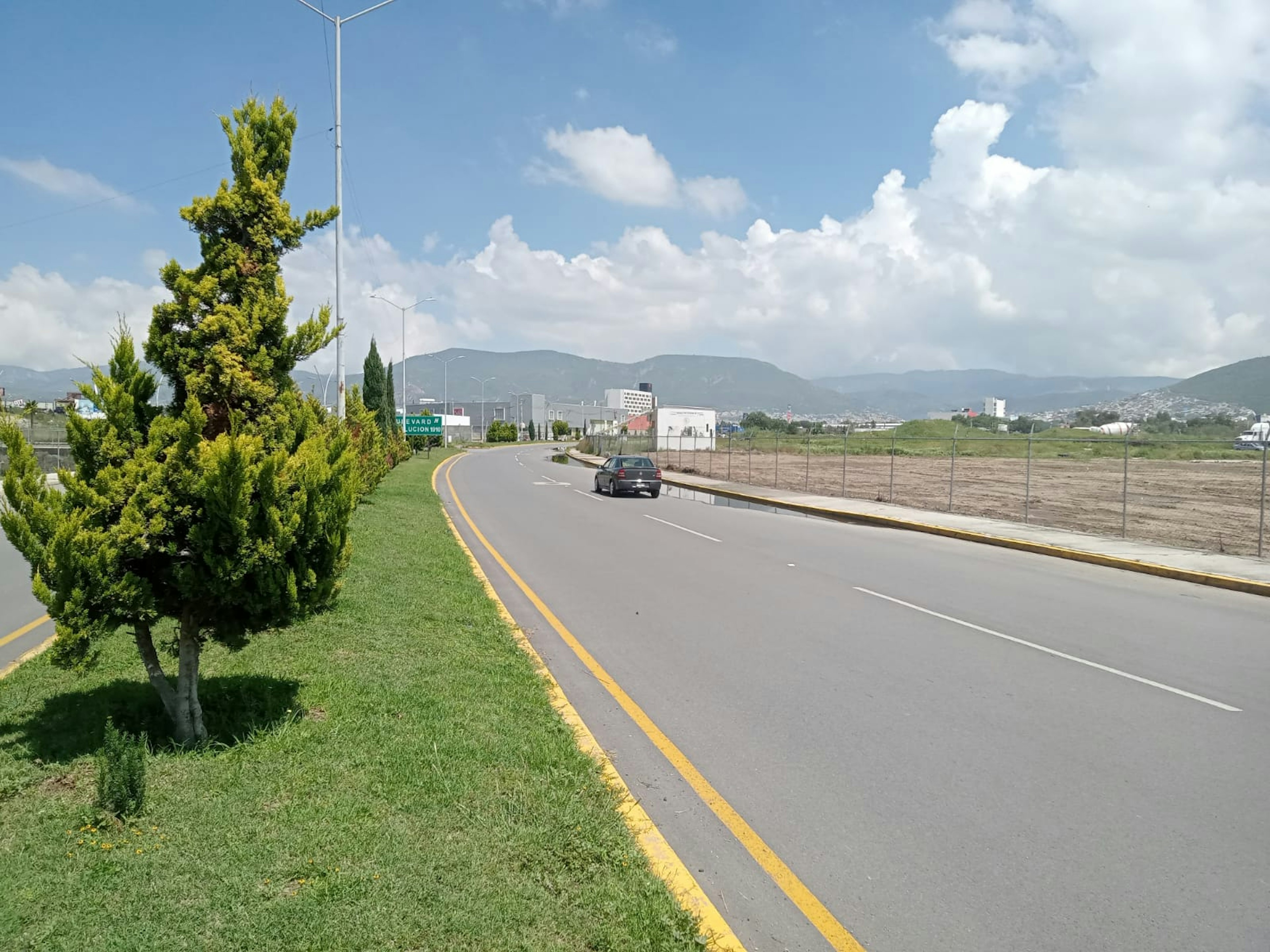 La imagen muestra una carretera amplia y bien mantenida que conduce a una zona urbana en desarrollo. A un lado se ve un área verde con árboles y arbustos cuidados. En el horizonte se observan montañas y edificios modernos, lo que sugiere una zona en expansión con potencial para nuevos desarrollos inmobiliarios. El cielo despejado y el entorno limpio indican un área atractiva para posibles inversiones en bienes raíces.