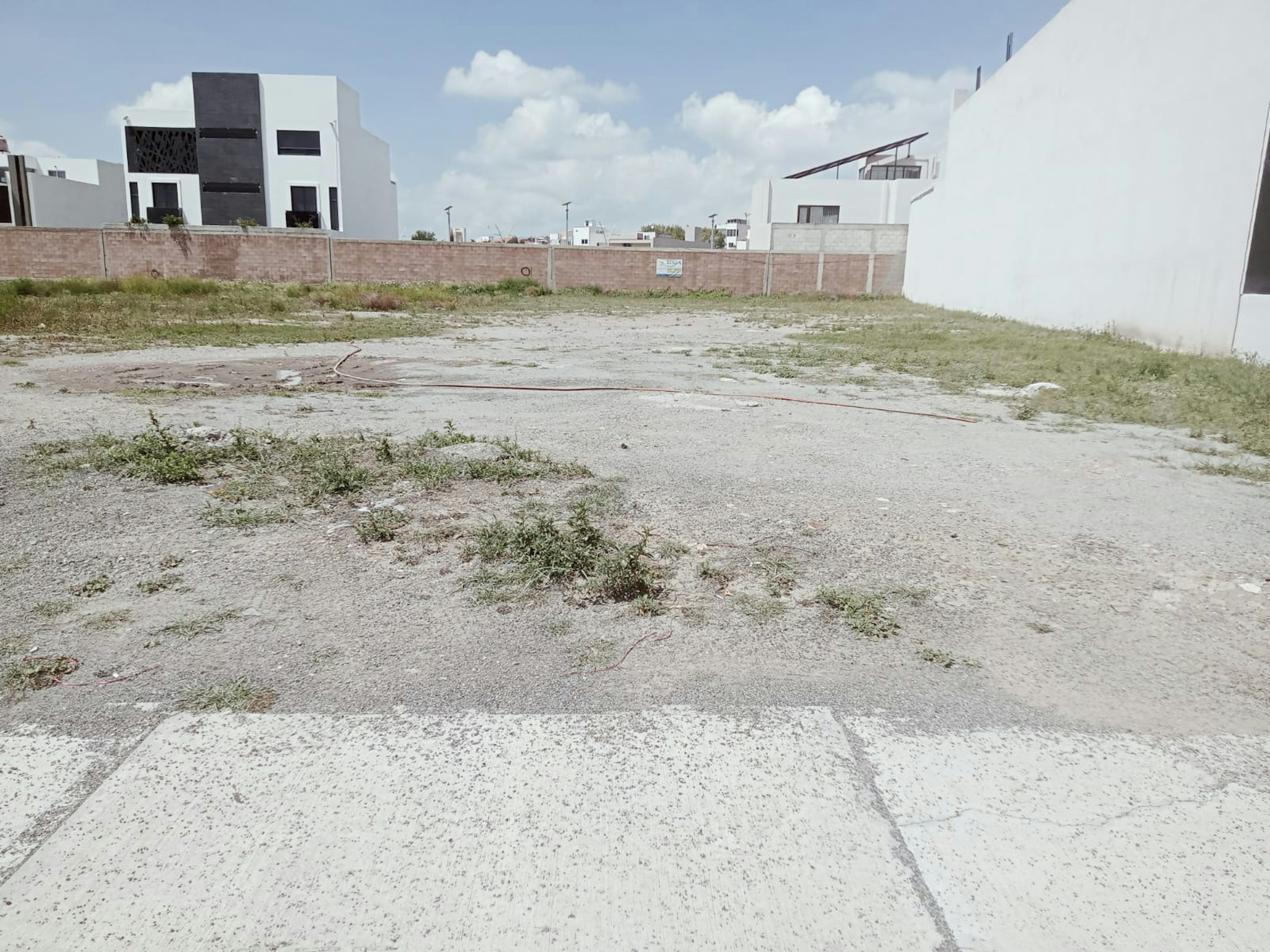 La imagen muestra un terreno baldío en una zona residencial en desarrollo. Se observa un lote vacío con vegetación escasa y suelo árido. Al fondo hay casas modernas de estilo contemporáneo y muros perimetrales. El cielo es azul con algunas nubes. El terreno parece estar listo para construcción, representando una oportunidad de inversión inmobiliaria en un área en crecimiento.