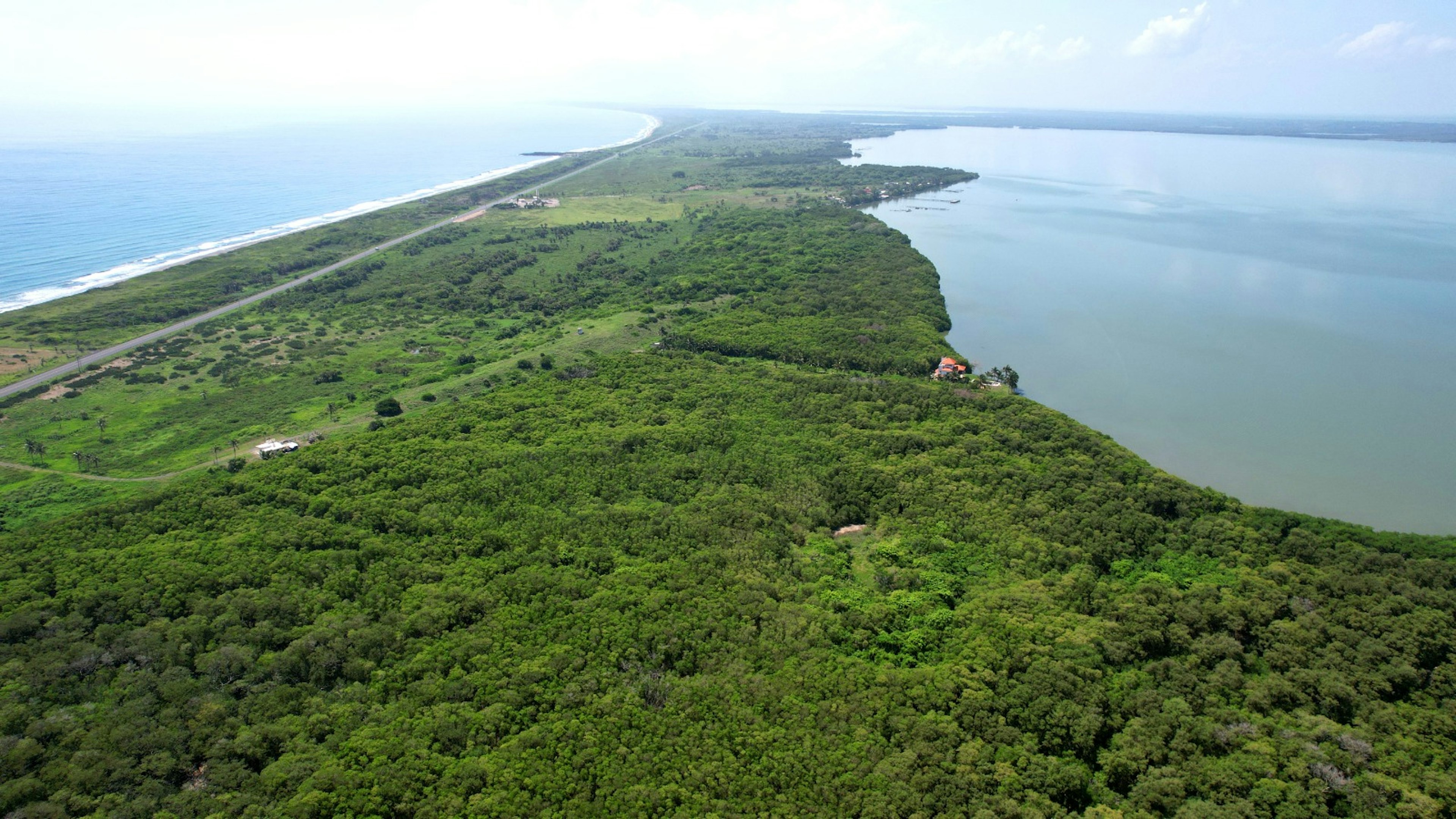 La imagen muestra una vista aérea de una zona costera tropical, con una extensa franja de tierra cubierta de vegetación densa. A un lado se observa una playa de arena blanca y el océano azul, mientras que al otro lado hay un cuerpo de agua más tranquilo, posiblemente una laguna o bahía. La zona parece ser un área natural protegida o poco desarrollada, ideal para proyectos ecoturísticos o resorts exclusivos que busquen integrar la naturaleza con el desarrollo inmobiliario sostenible.