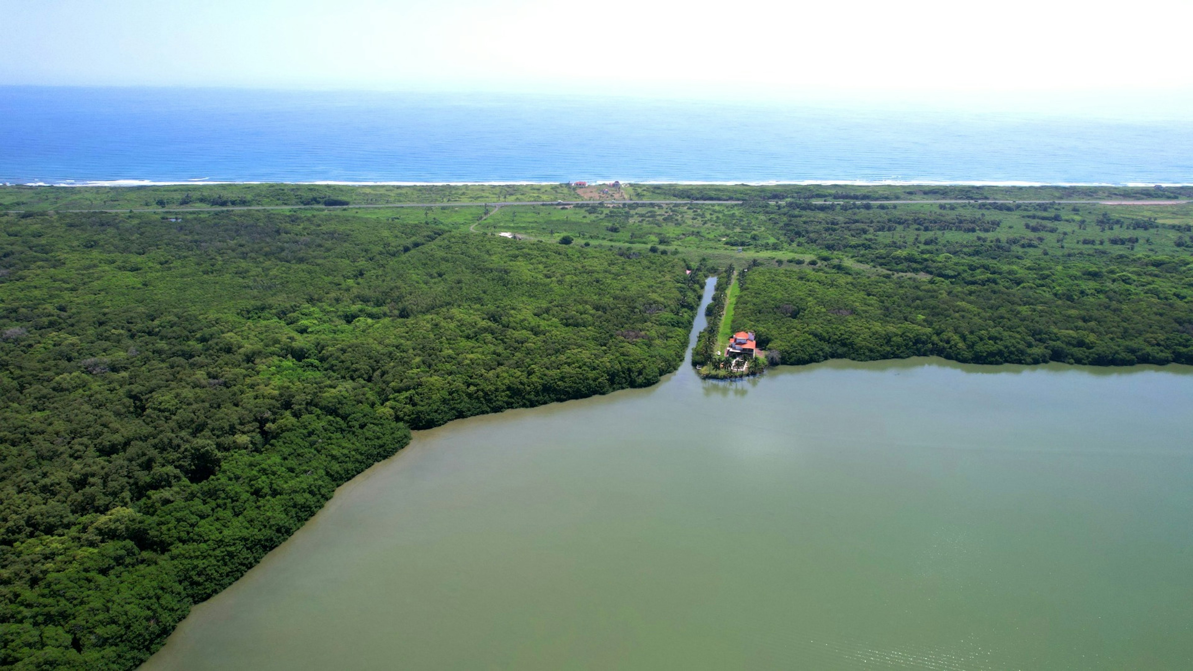 La imagen muestra una vista aérea de una zona costera con un extenso bosque verde y un gran cuerpo de agua. En el borde del agua, se observa una estructura con techo rojo, posiblemente una propiedad exclusiva. Un canal estrecho conecta la propiedad con el mar abierto visible en el horizonte. Esta ubicación ofrece una combinación única de privacidad, naturaleza y acceso al mar, ideal para un desarrollo inmobiliario de lujo o un resort ecológico.