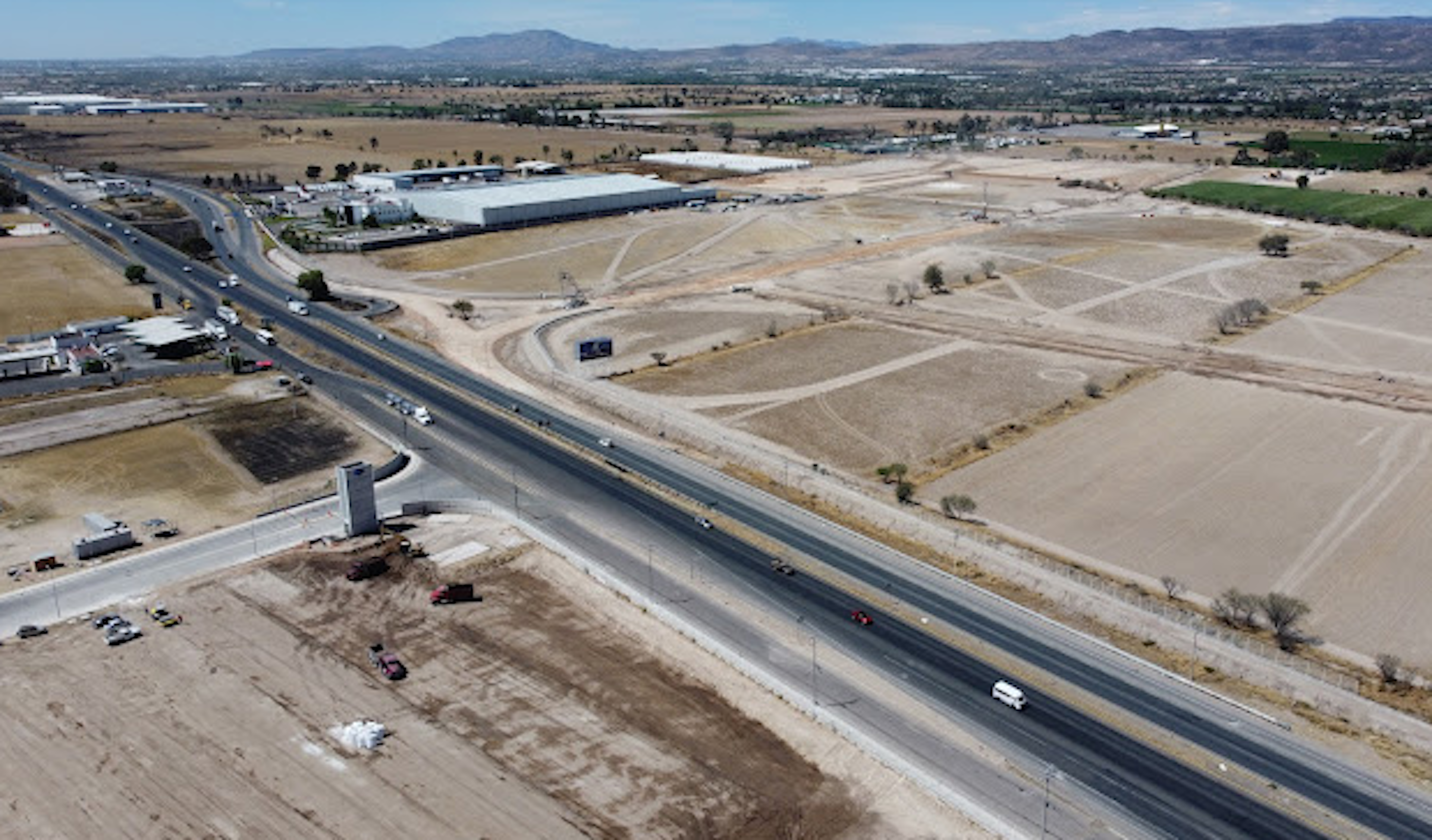 La imagen muestra una vista aérea de una zona industrial y comercial en desarrollo. Se observa una amplia carretera que atraviesa terrenos baldíos y áreas en construcción. Hay grandes edificios industriales y almacenes visibles, así como extensas áreas de terreno sin desarrollar. El paisaje es árido, con montañas en el fondo. Esta área parece estar en proceso de urbanización y expansión comercial, ofreciendo oportunidades para futuros desarrollos inmobiliarios.