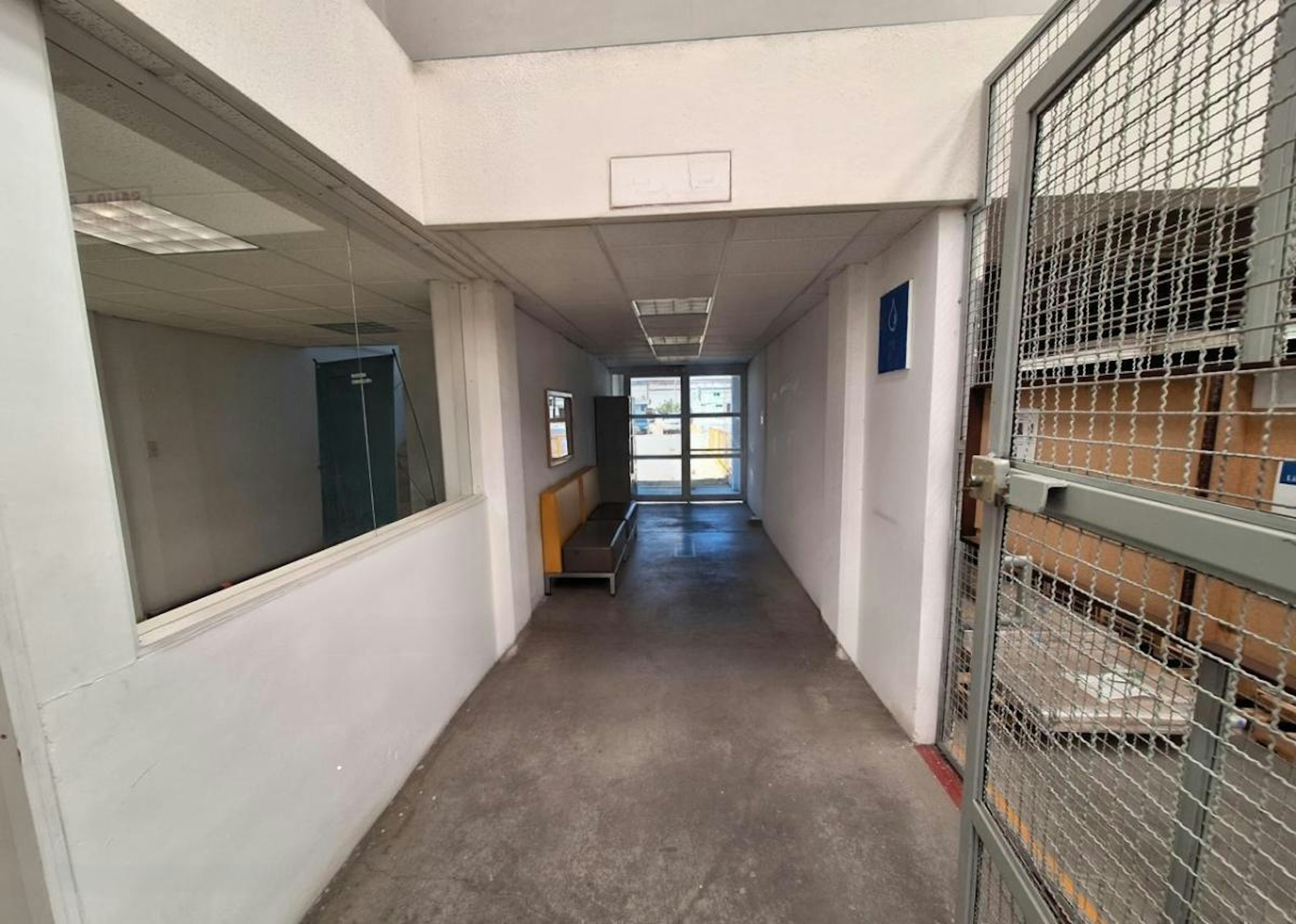La imagen muestra el interior de un edificio industrial o comercial. Se observa un pasillo con paredes blancas, un piso de concreto y una salida de emergencia señalizada en español. Hay una ventana con rejas metálicas a la derecha y una puerta de vidrio al fondo. El espacio parece estar en proceso de remodelación o acondicionamiento para uso industrial o logístico, como lo sugiere el texto superpuesto en la imagen.