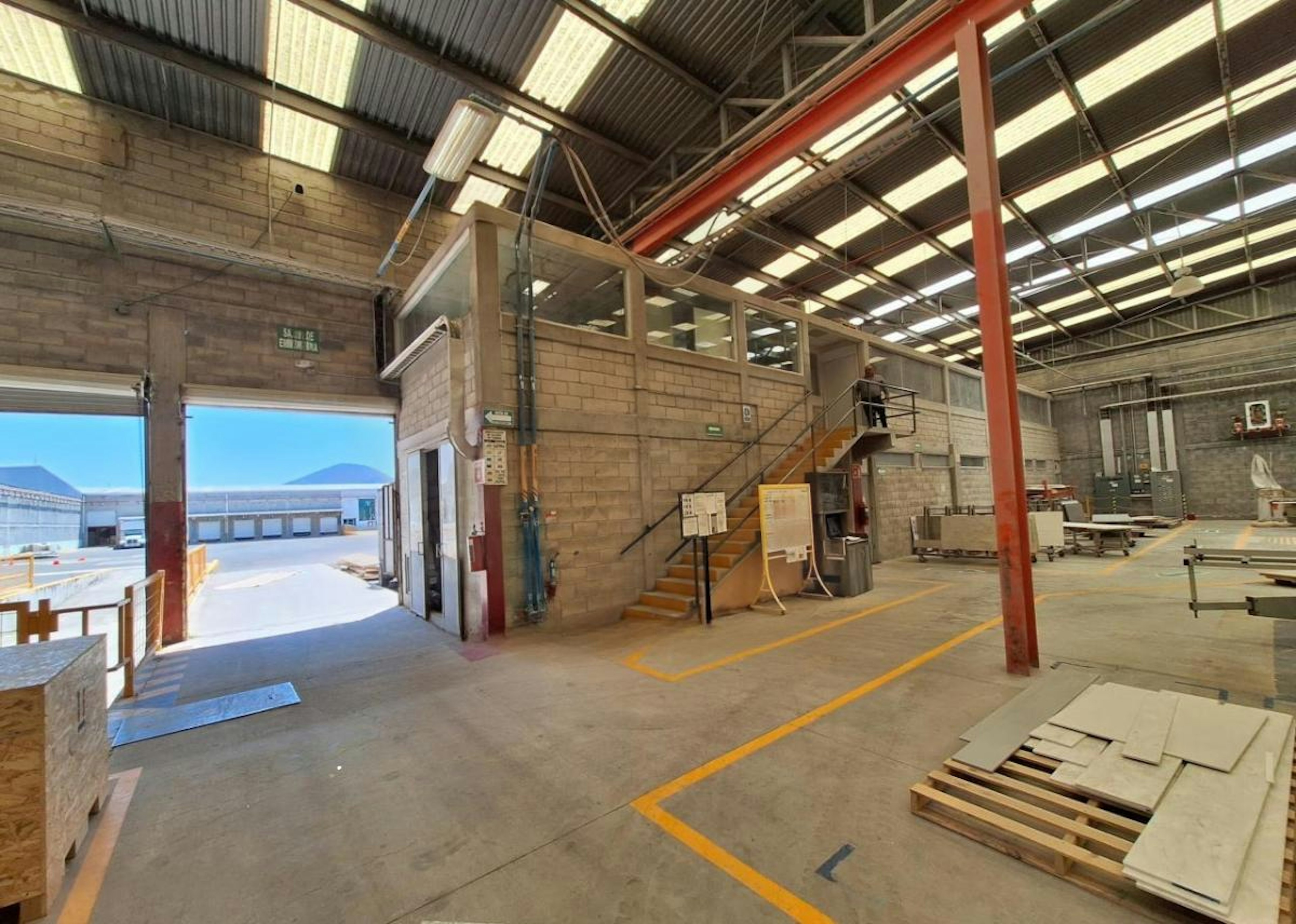 La imagen muestra el interior de una nave industrial o almacén logístico. Se observa una estructura de techo alto con vigas metálicas y tragaluces. Hay un área de oficinas construida dentro del espacio, con paredes de bloques y una escalera. Se ve una puerta grande abierta que da al exterior, donde se aprecian otras instalaciones industriales. El suelo tiene marcas de seguridad amarillas y hay algunos materiales de construcción dispersos.