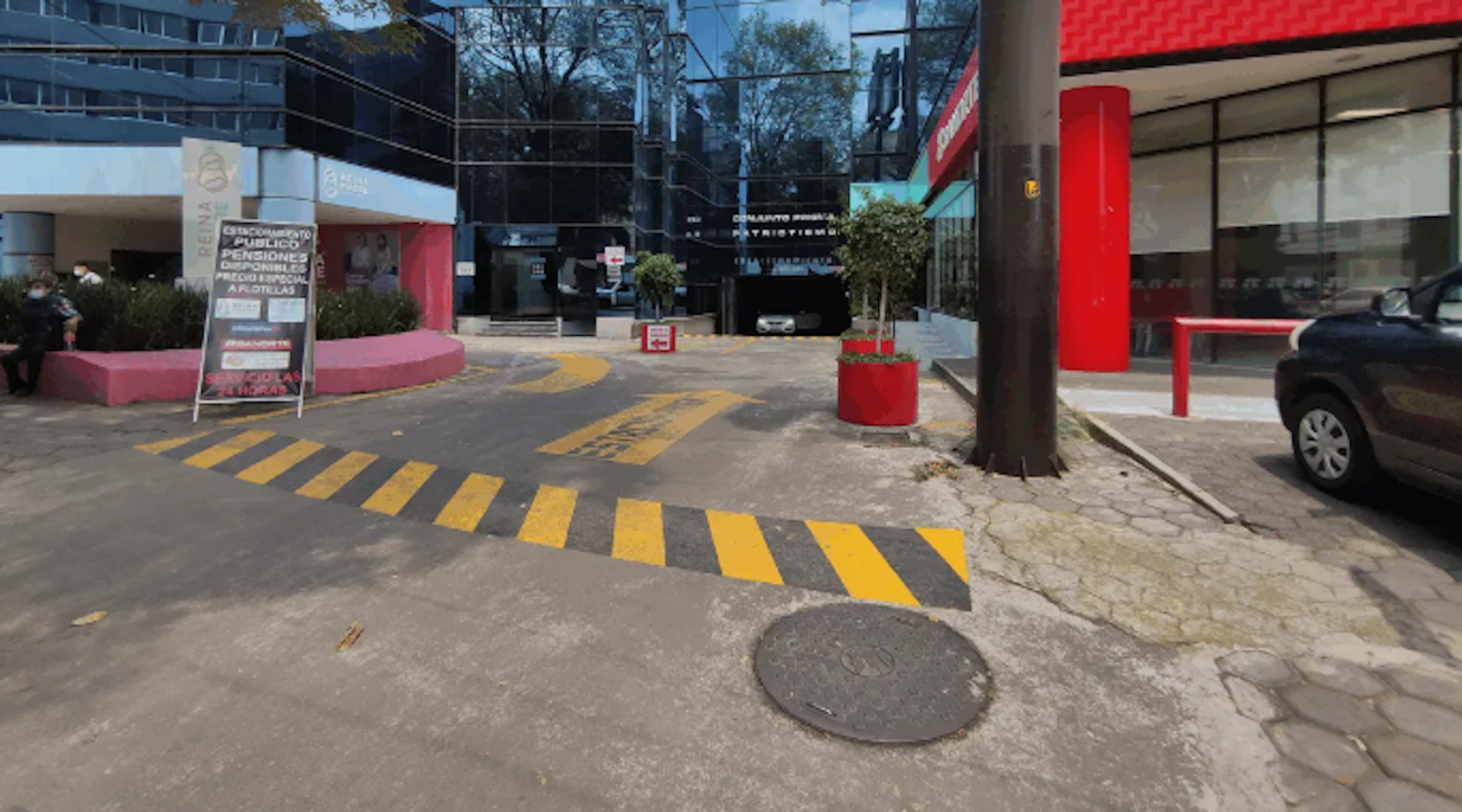 La imagen muestra la entrada a un estacionamiento público o área comercial. Se observa un camino con franjas amarillas de precaución, edificios modernos con fachadas de vidrio, y elementos decorativos como macetas rojas. Hay señalización que indica servicios públicos y un ambiente urbano bien mantenido. La escena sugiere una zona comercial o de oficinas con instalaciones para vehículos.