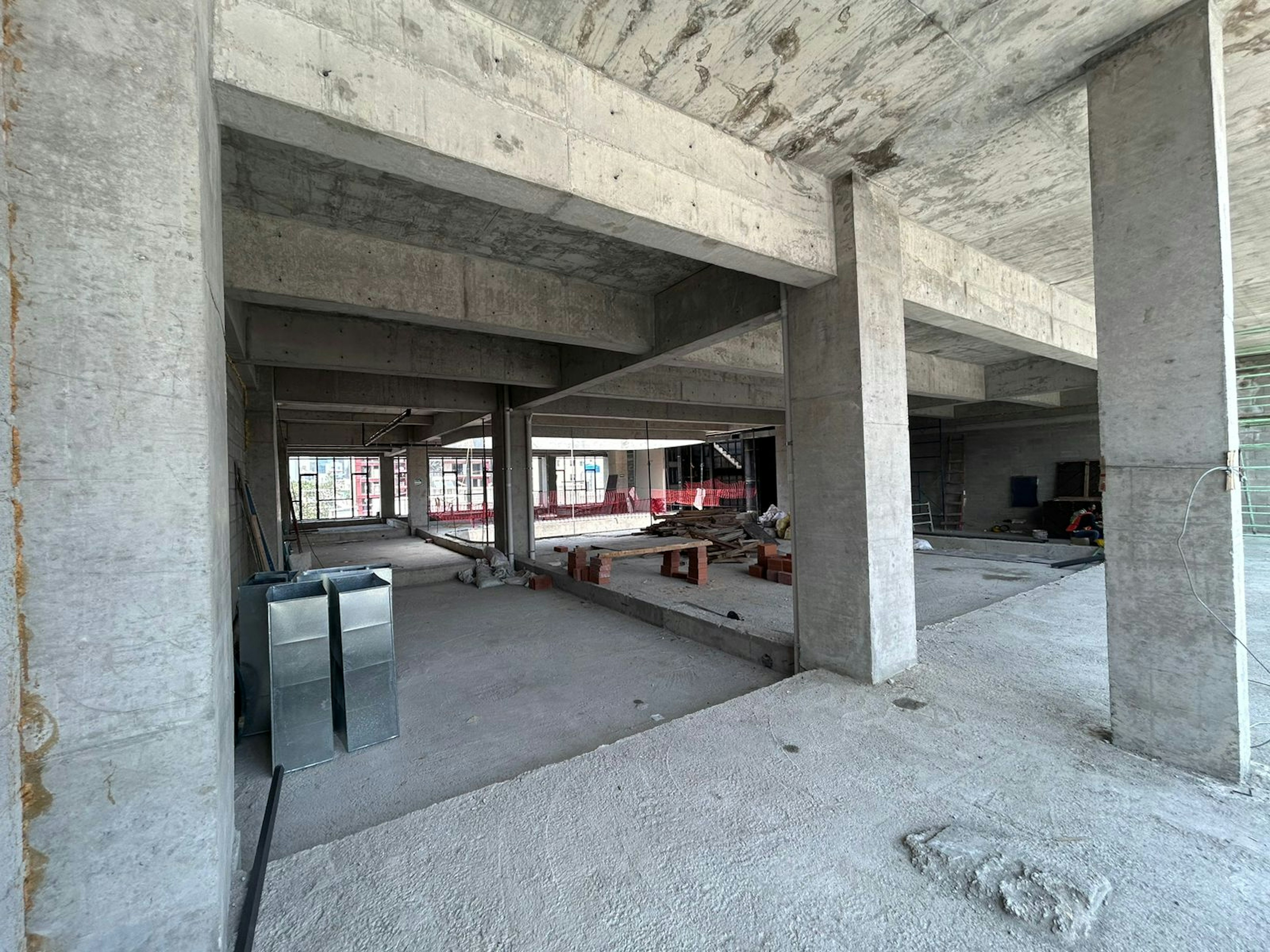 Interior de una construcción en proceso, con columnas de concreto y espacios abiertos sin acabados. Materiales de construcción y herramientas dispersos en el suelo.