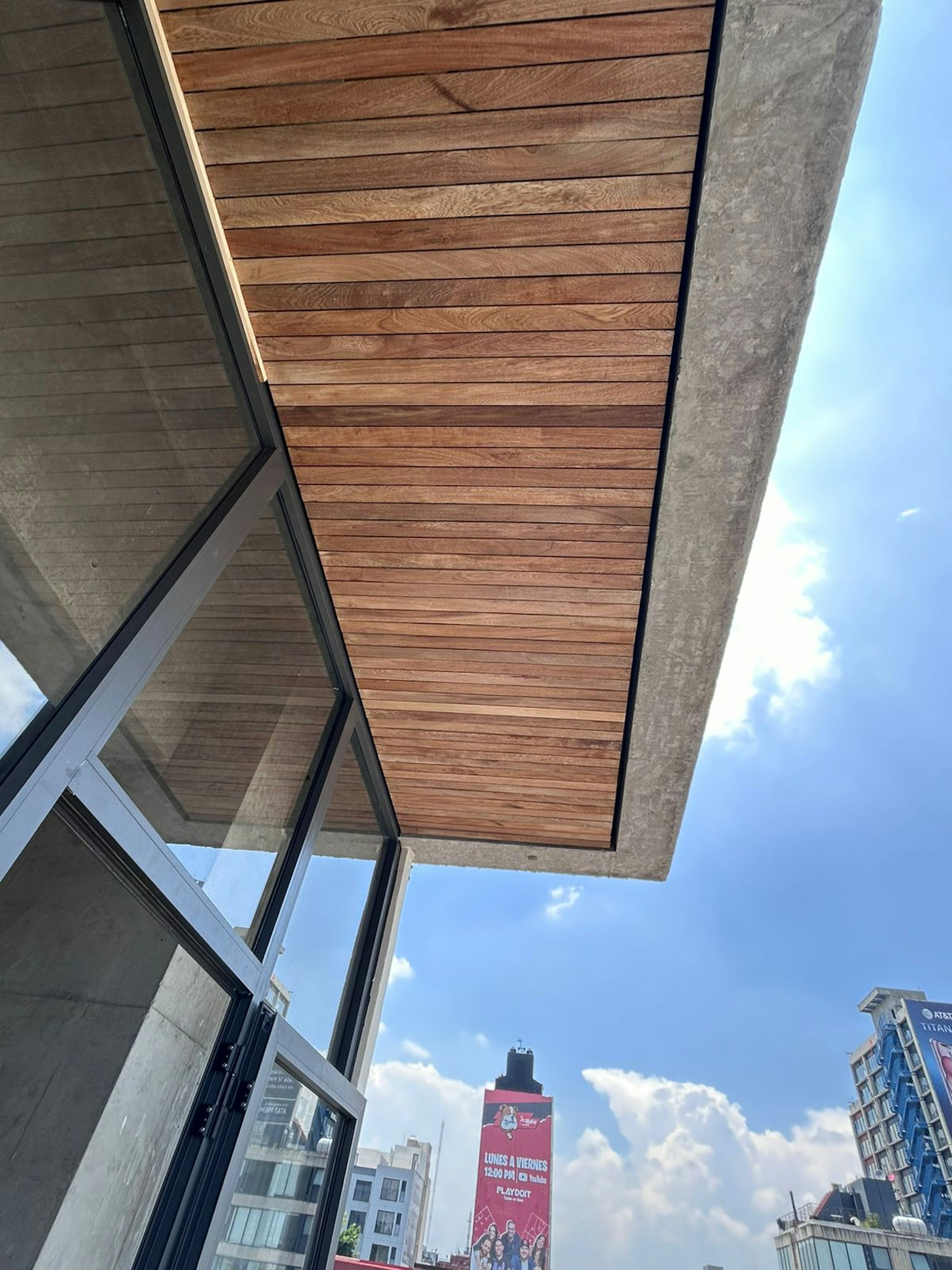 La imagen muestra una perspectiva arquitectónica desde el interior de un edificio moderno, destacando materiales como vidrio y madera, con vistas a edificios urbanos y un cielo despejado. Ideal para promocionar espacios abiertos y diseño contemporáneo en el sector inmobiliario.