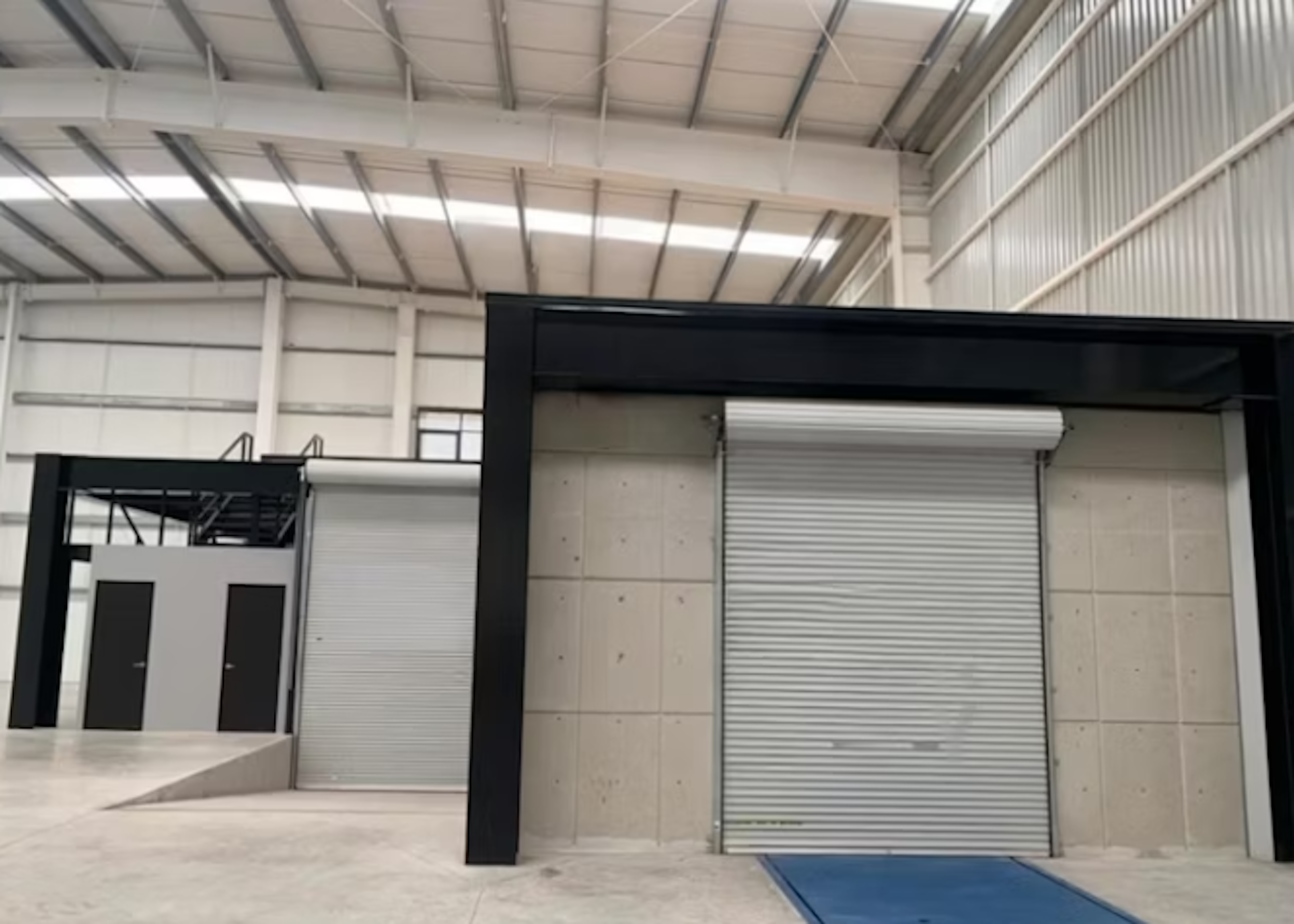 La imagen muestra el interior de un edificio industrial o almacén moderno. Se observa una estructura de techo alta con vigas expuestas, paredes blancas y un piso de concreto. Hay una puerta enrollable grande y una estructura negra que parece ser un marco para otra puerta o área. A la izquierda se ve una pequeña plataforma elevada con escaleras y dos puertas. El espacio parece estar en buenas condiciones y listo para uso comercial o industrial.