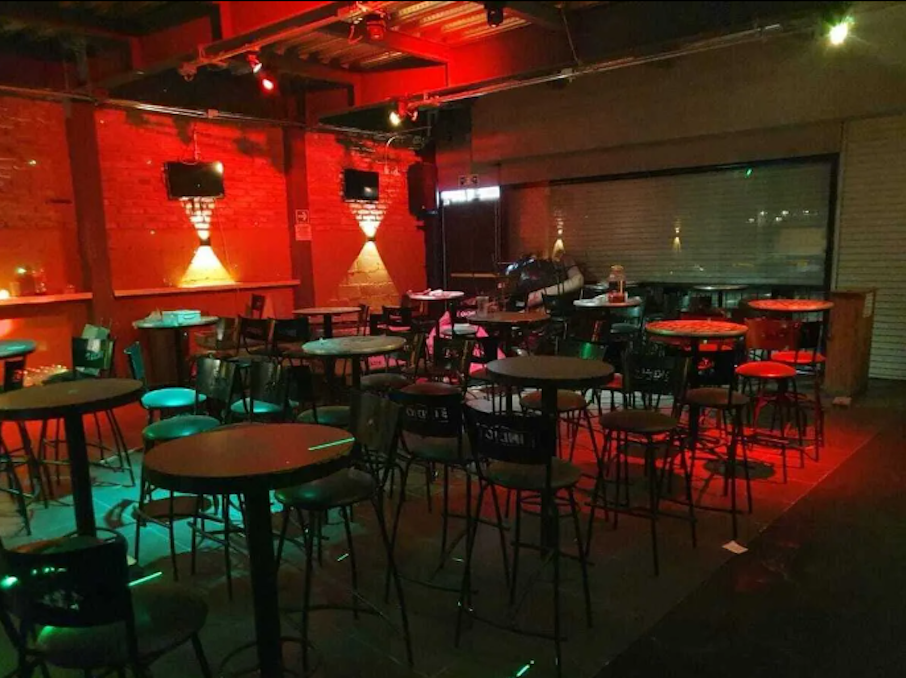 Este local parece ser un bar o club nocturno con un ambiente íntimo y moderno. El espacio cuenta con mesas altas y taburetes, iluminación roja y verde que crea un ambiente vibrante. Las paredes de ladrillo rojo, los focos direccionales y las pantallas en la pared añaden un toque industrial y contemporáneo. El local está vacío, sugiriendo que podría estar cerrado o preparándose para abrir.