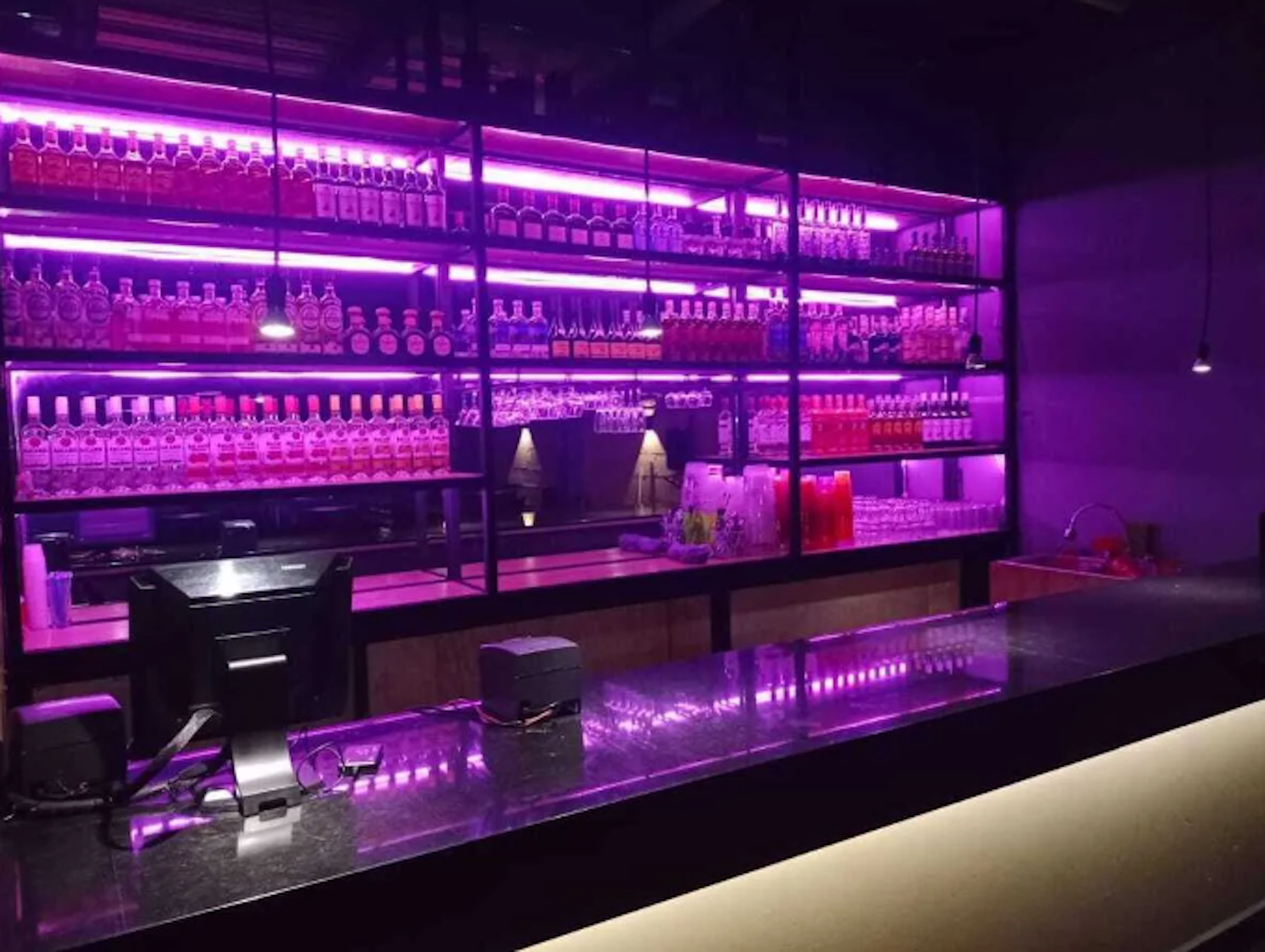 Esta imagen muestra un bar o club nocturno moderno y elegante. La barra está iluminada con luces LED púrpuras, creando una atmósfera vibrante y de alta energía. Los estantes detrás de la barra están llenos de botellas de licor, organizadas cuidadosamente y también iluminadas. El diseño es contemporáneo, con líneas limpias y un ambiente sofisticado, ideal para un espacio de entretenimiento en un edificio comercial o residencial de lujo.