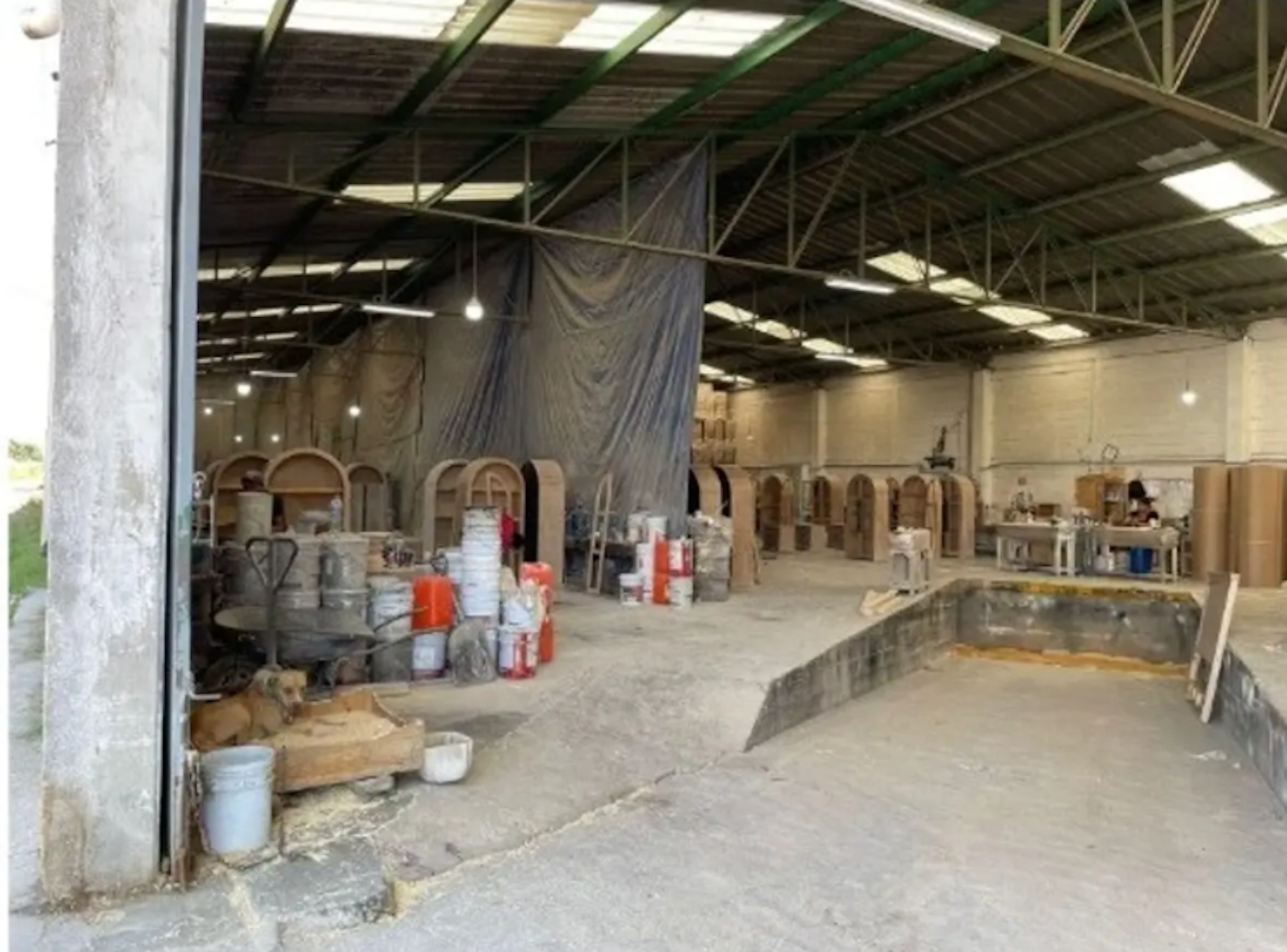 Esta imagen muestra un amplio espacio industrial en construcción o renovación. Se observa una estructura metálica en el techo, paredes parcialmente terminadas y diversos materiales de construcción dispersos por el suelo. Hay arcos decorativos apoyados contra la pared, sugiriendo un posible uso comercial o residencial futuro. Un perro descansa en una esquina, añadiendo un toque inesperado a la escena de obra.