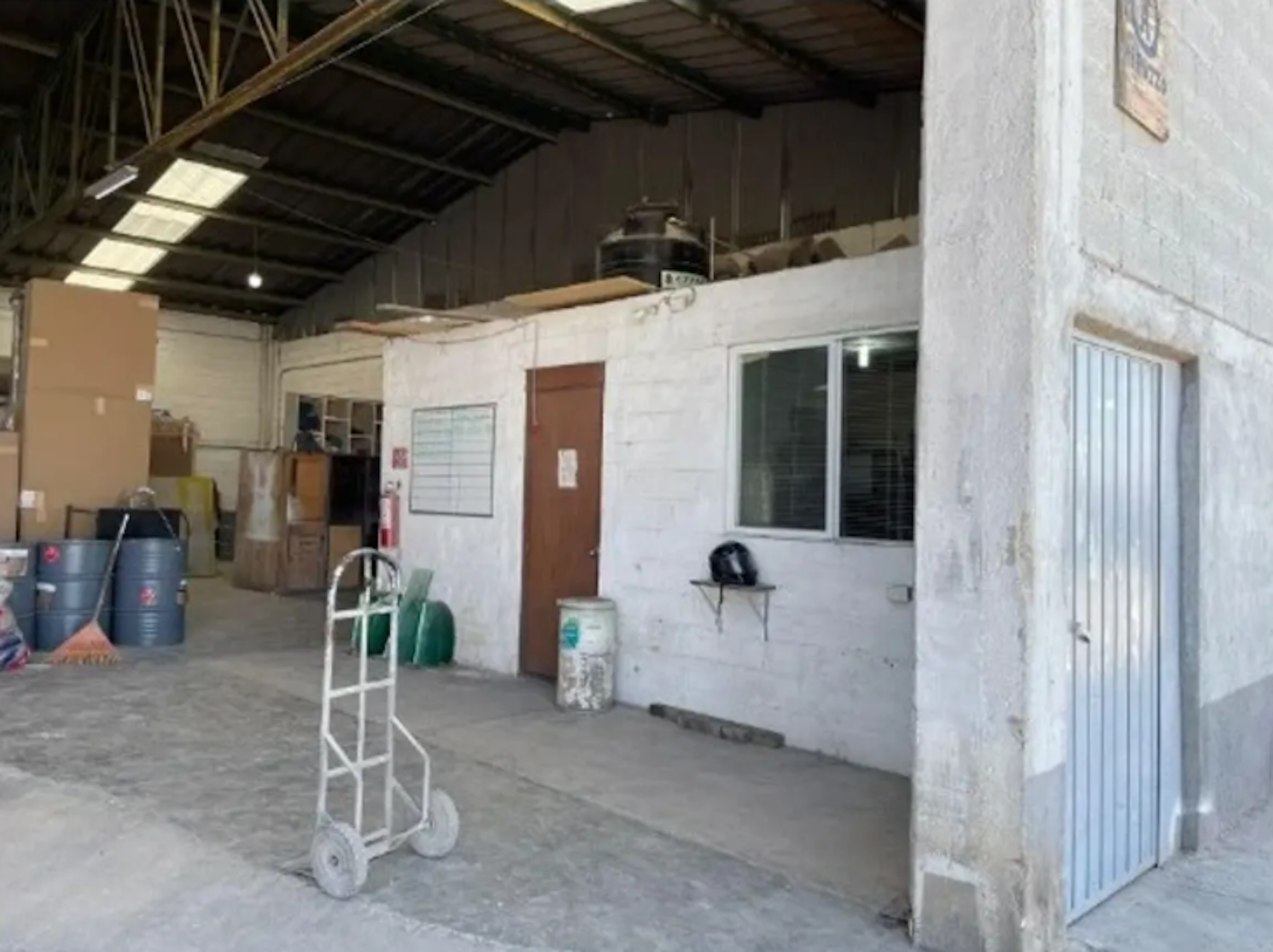 La imagen muestra el interior de un almacén o bodega industrial con techo alto. Se ve una oficina o área administrativa construida dentro del espacio, con paredes blancas, una puerta marrón y una ventana. Hay varios elementos de almacenamiento como barriles y un carro de carga. El espacio parece estar en uso pero algo desordenado, típico de un ambiente de trabajo industrial o de logística.