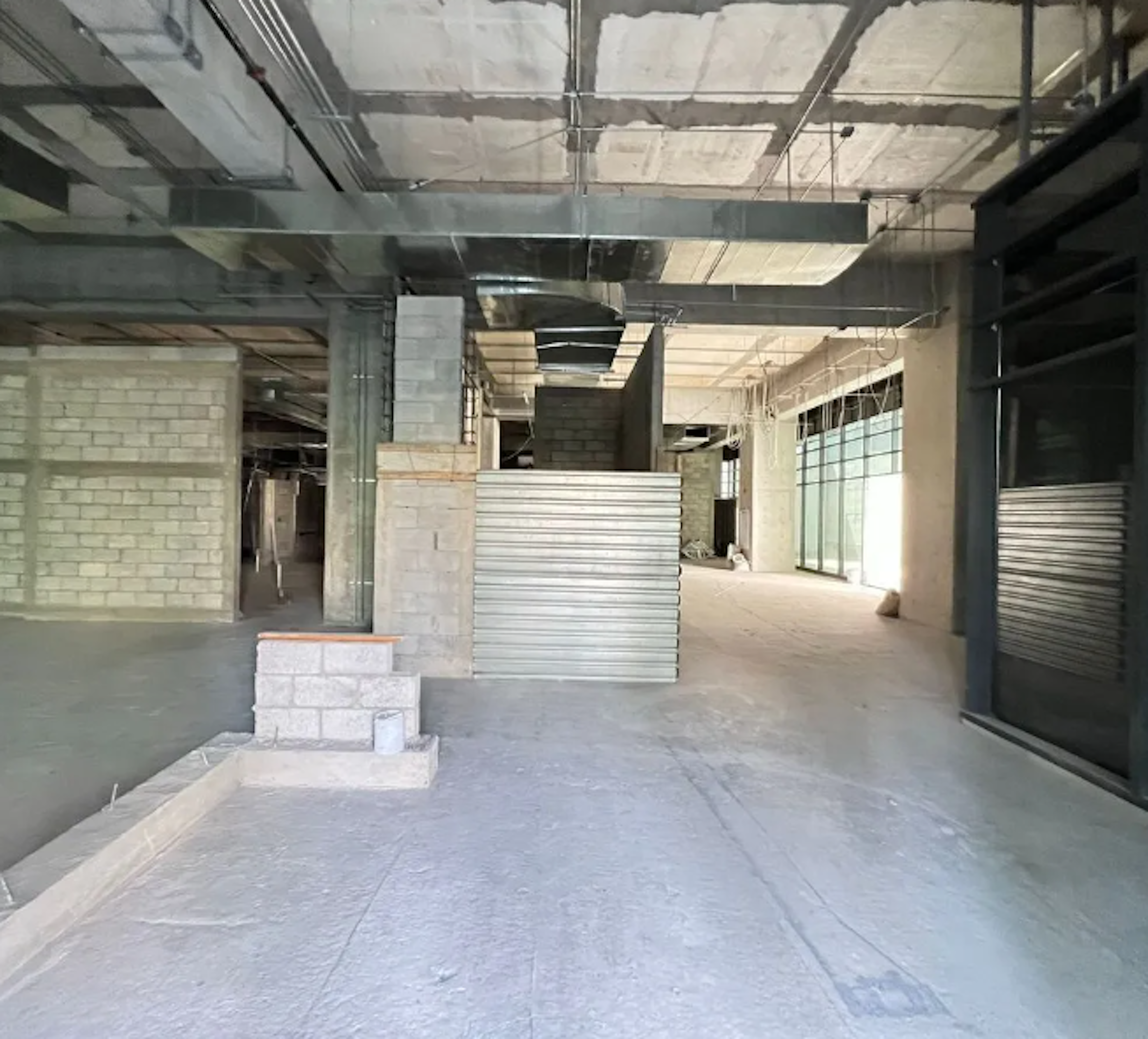 La imagen muestra un espacio comercial en construcción o renovación. Se observa un interior amplio y sin terminar, con paredes de ladrillo expuesto, techos altos con conductos visibles, y pisos de concreto. Hay áreas parcialmente construidas y materiales de construcción dispersos. La iluminación natural entra por ventanas grandes, sugiriendo un potencial diseño moderno e industrial para el espacio una vez finalizado.