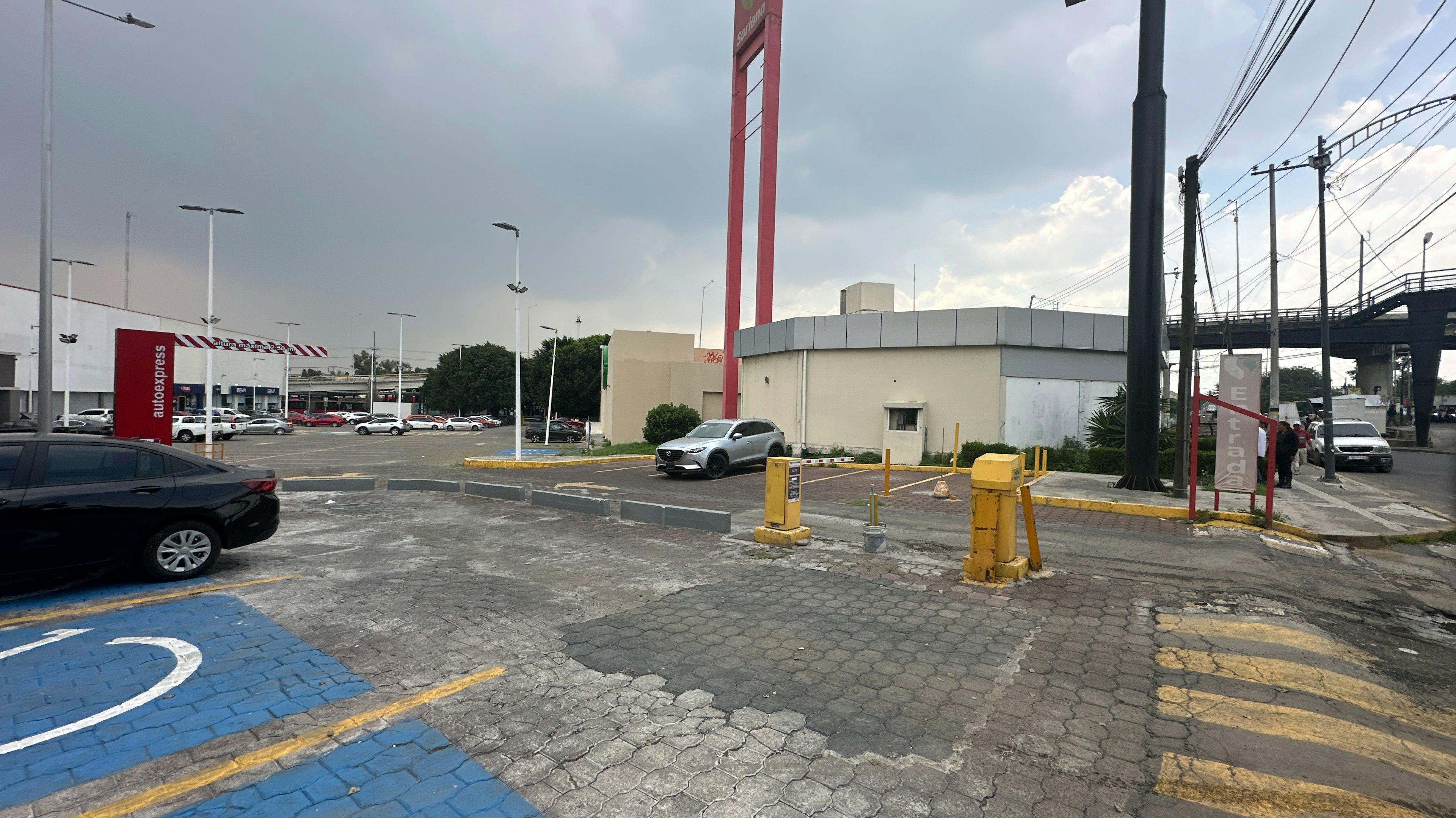 La imagen muestra un área comercial con un estacionamiento y varios edificios. Se observa una estructura con una torre roja alta, posiblemente parte de un centro comercial o tienda departamental. Hay vehículos estacionados y en movimiento, así como infraestructura urbana como postes de luz y señalización vial. El pavimento muestra marcas de estacionamiento y el cielo está parcialmente nublado.