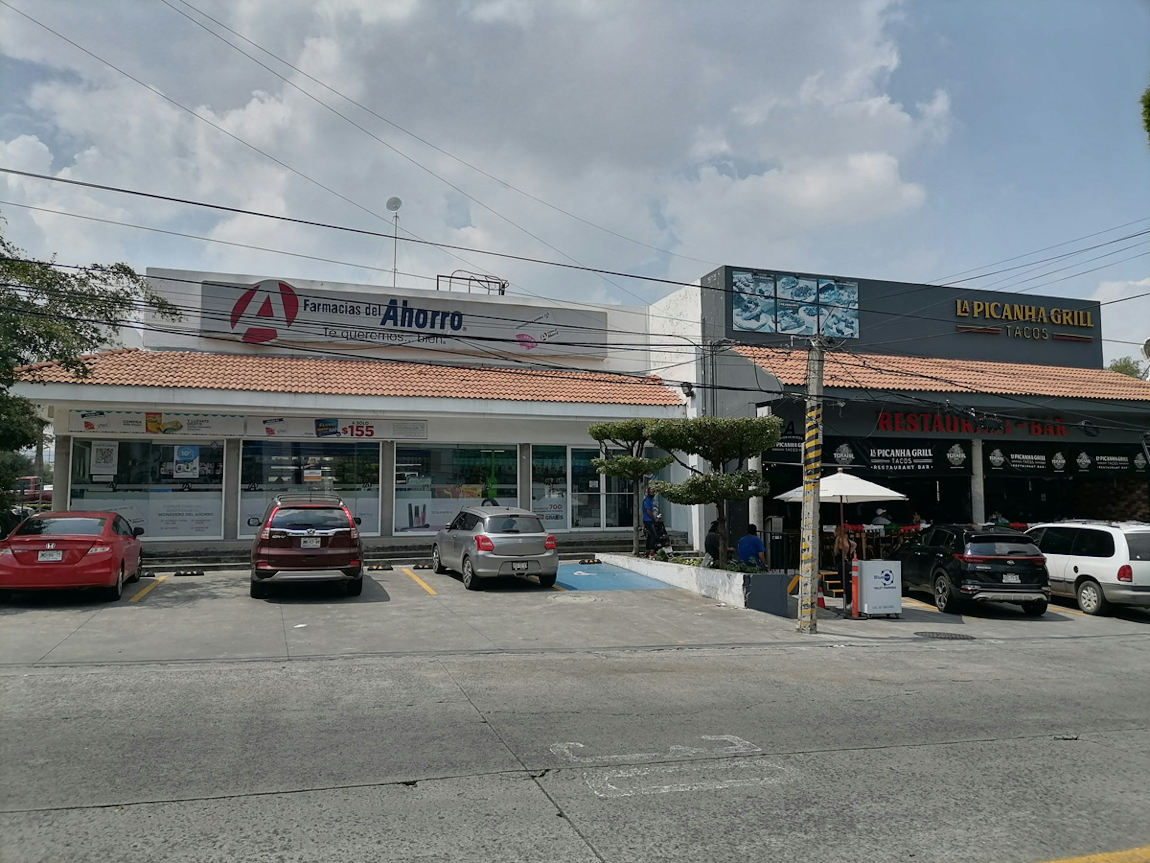 La imagen muestra un área comercial con dos establecimientos principales: una farmacia llamada Farmacias del Ahorro y un restaurante de tacos llamado La Picanha Grill. El edificio tiene un estilo arquitectónico mixto, con techos de tejas y fachadas modernas. Hay un estacionamiento en frente con varios vehículos aparcados. La escena sugiere una zona suburbana o de pequeña ciudad con una mezcla de negocios locales.