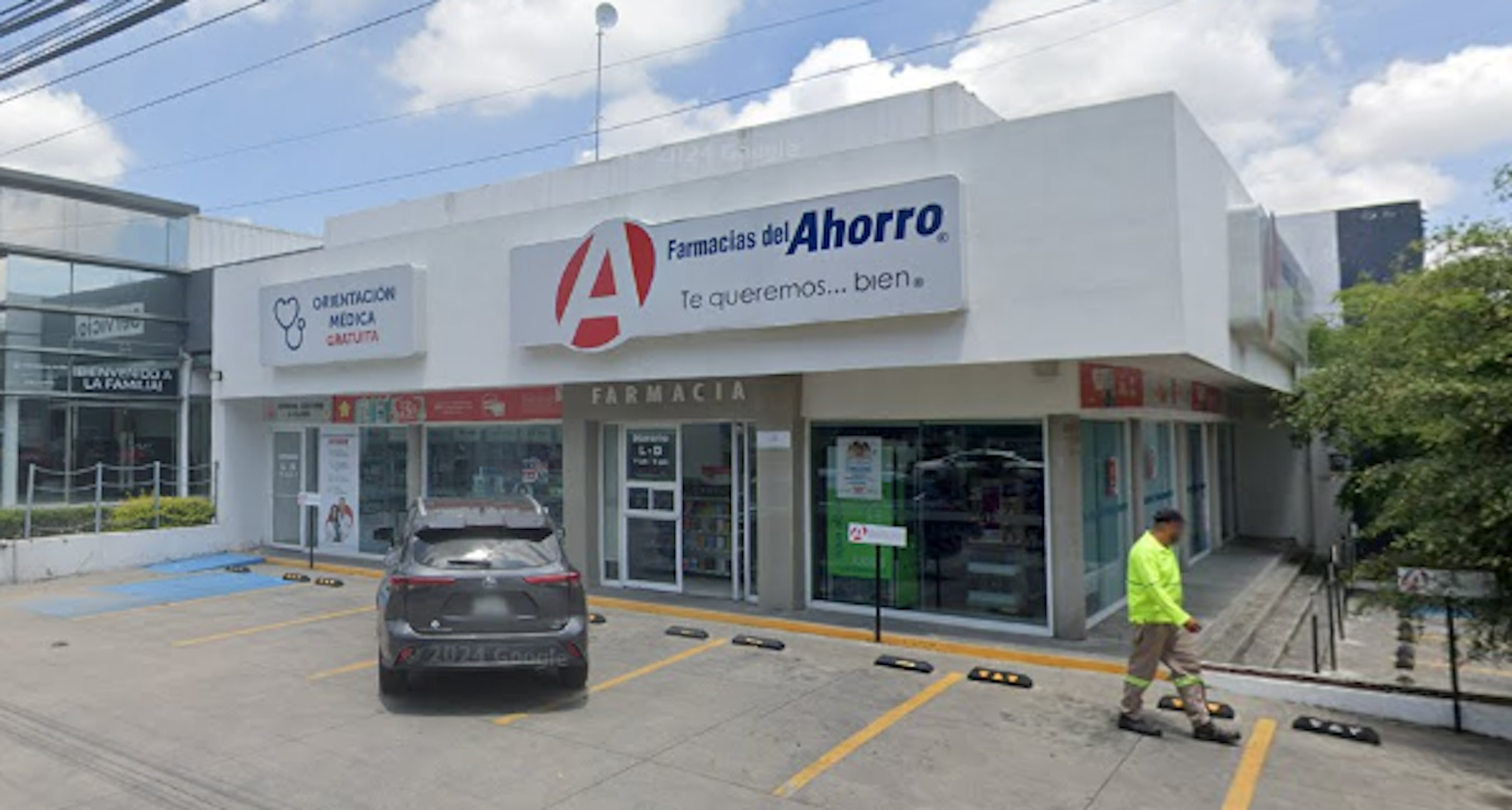 La imagen muestra una farmacia llamada Farmacias del Ahorro ubicada en una zona comercial. El edificio es de color blanco con un letrero prominente. Hay un estacionamiento en frente con un automóvil estacionado. La fachada tiene ventanas grandes y puertas de vidrio. Se observa también un letrero de orientación médica gratuita. El entorno sugiere una ubicación en una zona urbana con cables eléctricos visibles.