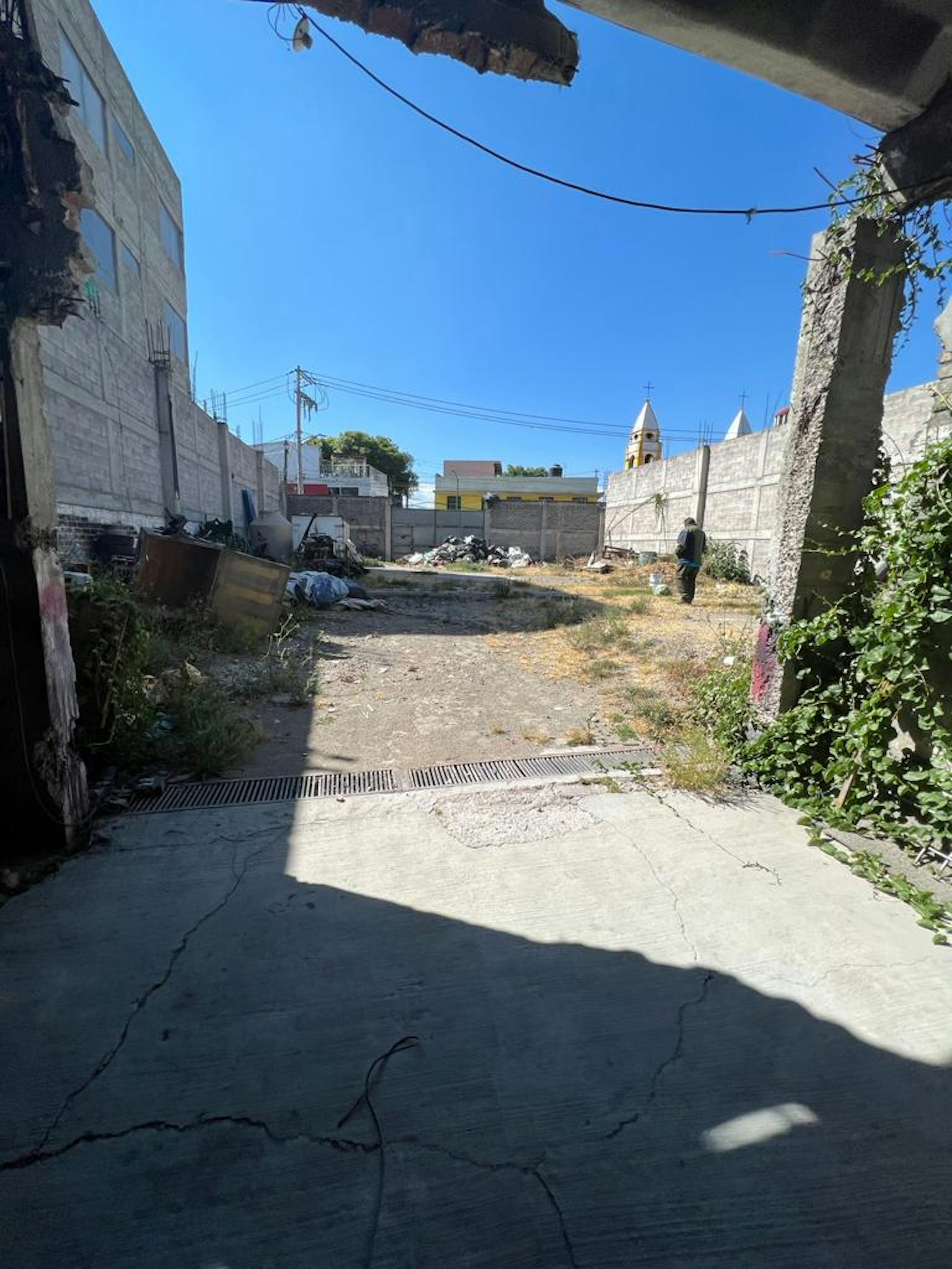 La imagen muestra un terreno baldío en una zona urbana. El área está rodeada de muros de concreto y edificios inacabados. Se observa basura y escombros esparcidos, así como vegetación descuidada. Al fondo se ven torres de iglesia y cables eléctricos. El terreno parece abandonado y en mal estado, pero podría tener potencial para desarrollo inmobiliario si se limpia y acondiciona adecuadamente.