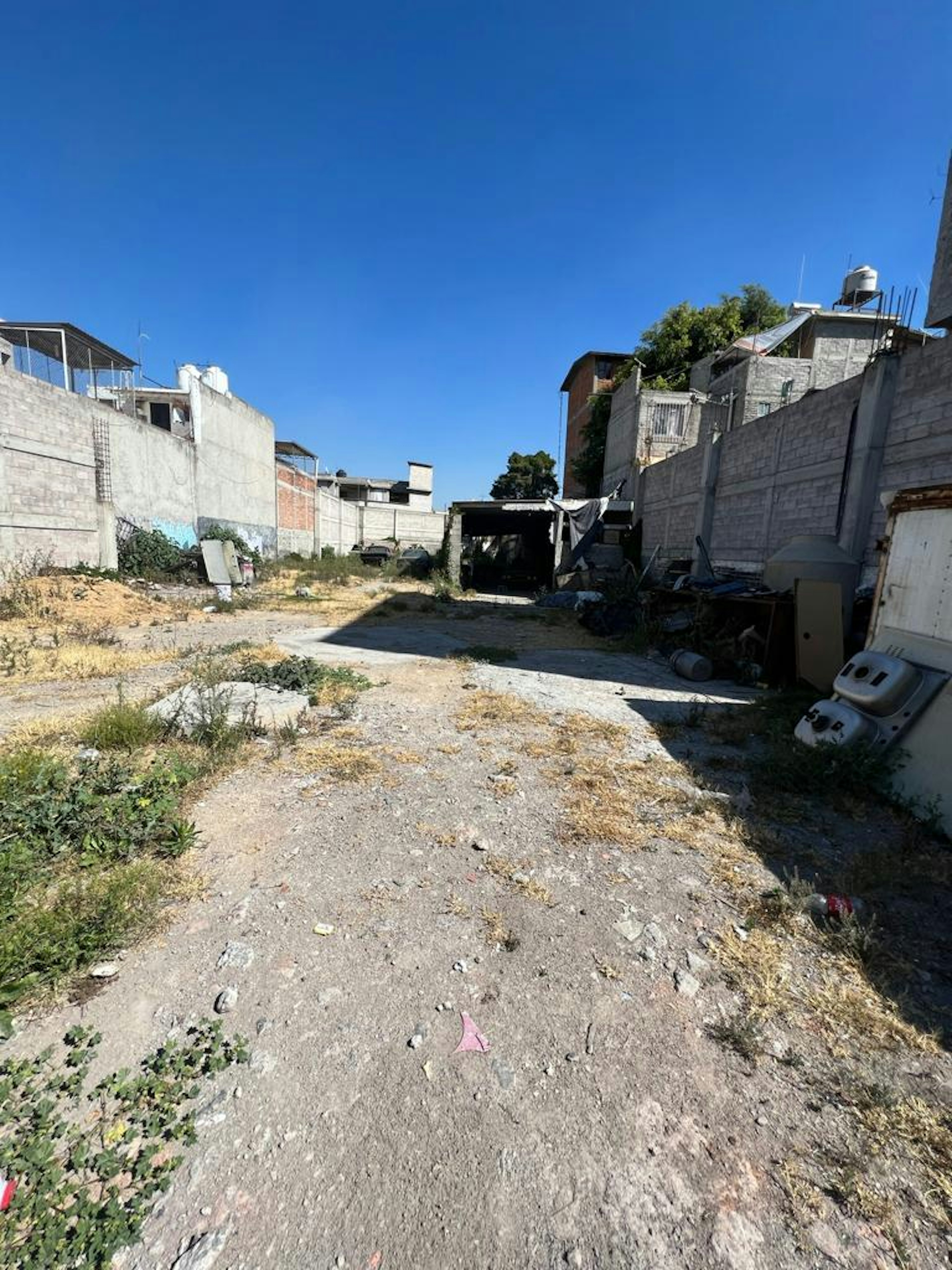 La imagen muestra un terreno baldío en una zona urbana de bajos recursos. Se observan muros de concreto deteriorados, escombros y vegetación dispersa. El área parece abandonada y descuidada, con basura y objetos desechados. Los edificios circundantes sugieren una zona residencial de construcción informal. El cielo azul contrasta con el estado ruinoso del terreno, que podría ser objeto de desarrollo o rehabilitación urbana.