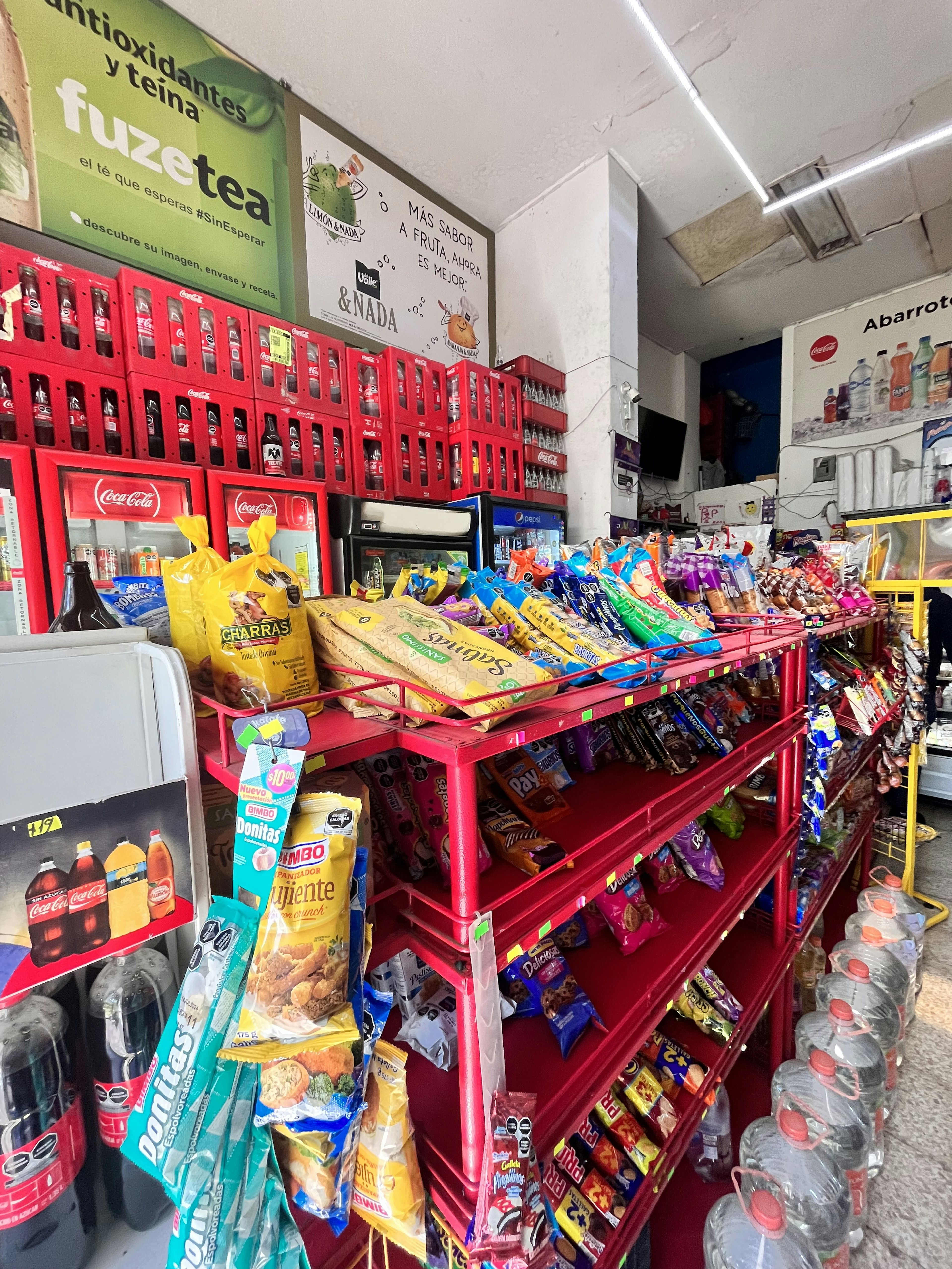 Interior de una tienda de abarrotes con estanterías llenas de productos variados como botanas y bebidas. Publicidad de marcas visibles y refrigerador.