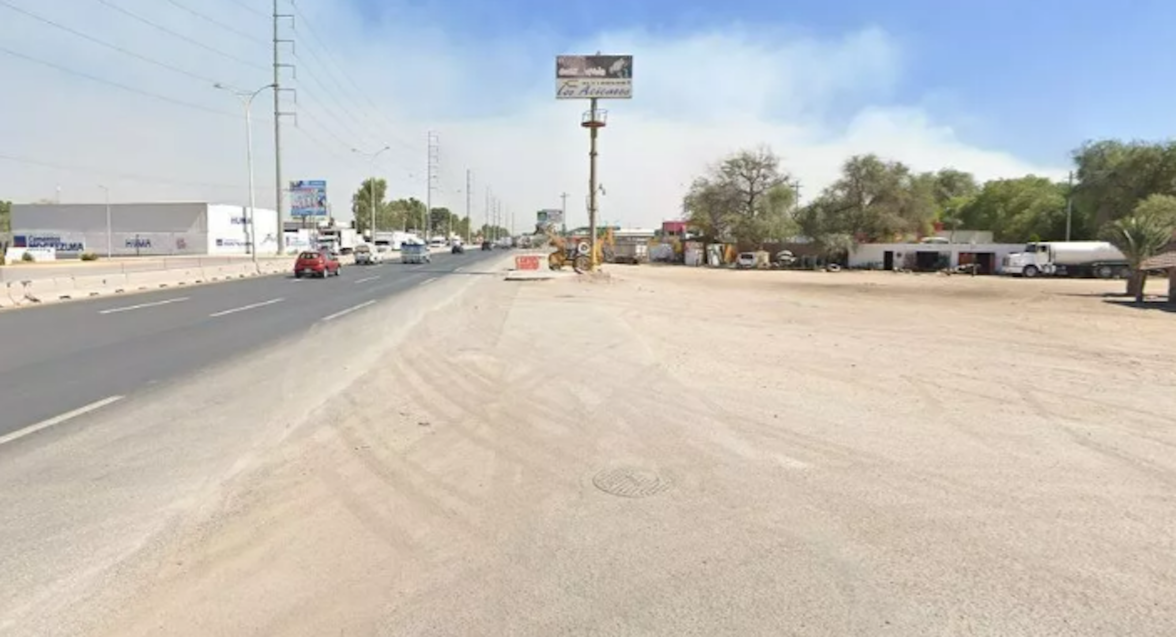 La imagen muestra una carretera amplia en una zona semi-urbana. A la izquierda se observa un edificio comercial y a la derecha un área sin desarrollar con algunos vehículos estacionados. Hay señalizaciones y postes de luz a lo largo de la vía. El entorno sugiere una zona en desarrollo con potencial para futuros proyectos inmobiliarios o comerciales, dada la infraestructura existente y el espacio disponible.