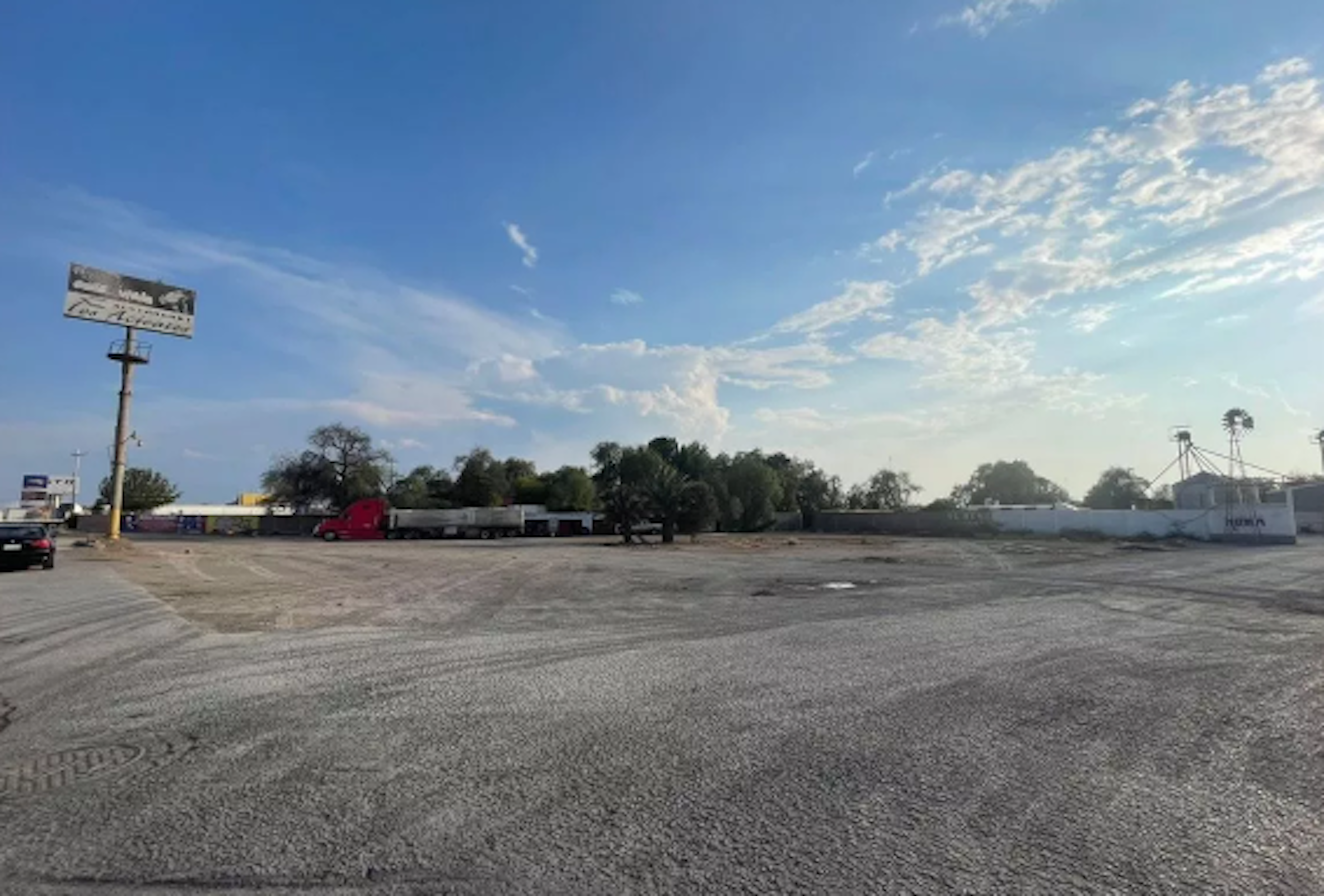 La imagen muestra un terreno amplio y plano, posiblemente un lote comercial o industrial. El área está mayormente sin desarrollar, con una superficie de grava o tierra compactada. Se observan algunos vehículos y estructuras en la distancia, así como árboles y vegetación en el fondo. Un letrero publicitario y postes eléctricos indican cierta infraestructura. El cielo azul y despejado sugiere un clima favorable para el desarrollo.