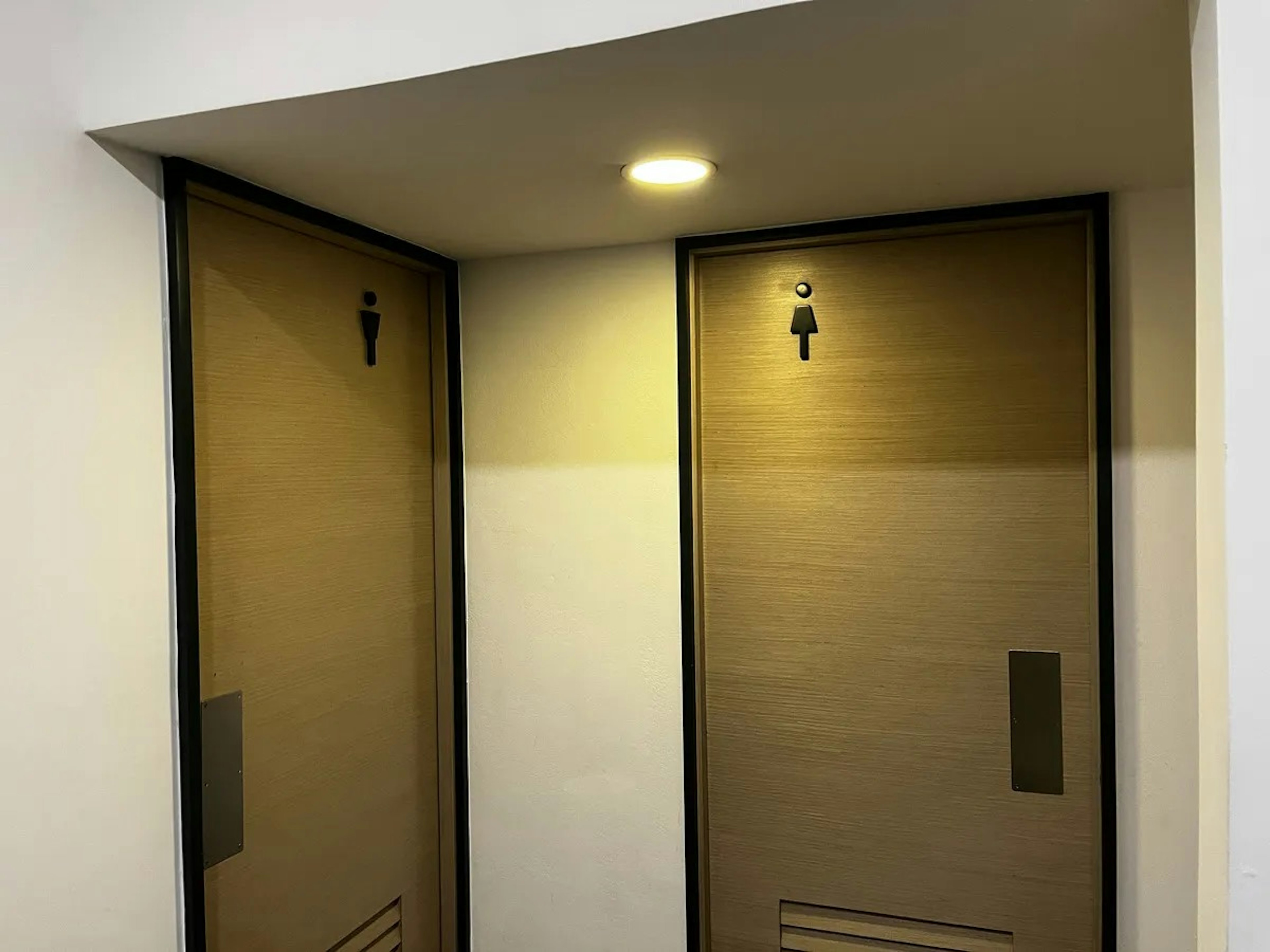 La imagen muestra dos puertas de baño públicos con acabados modernos. Las puertas son de color beige claro con marcos negros. Cada puerta tiene un icono que indica si es para hombres o mujeres. Hay una luz en el techo que ilumina el área, creando un ambiente cálido. El diseño es minimalista y funcional, típico de instalaciones comerciales o de oficinas actuales.