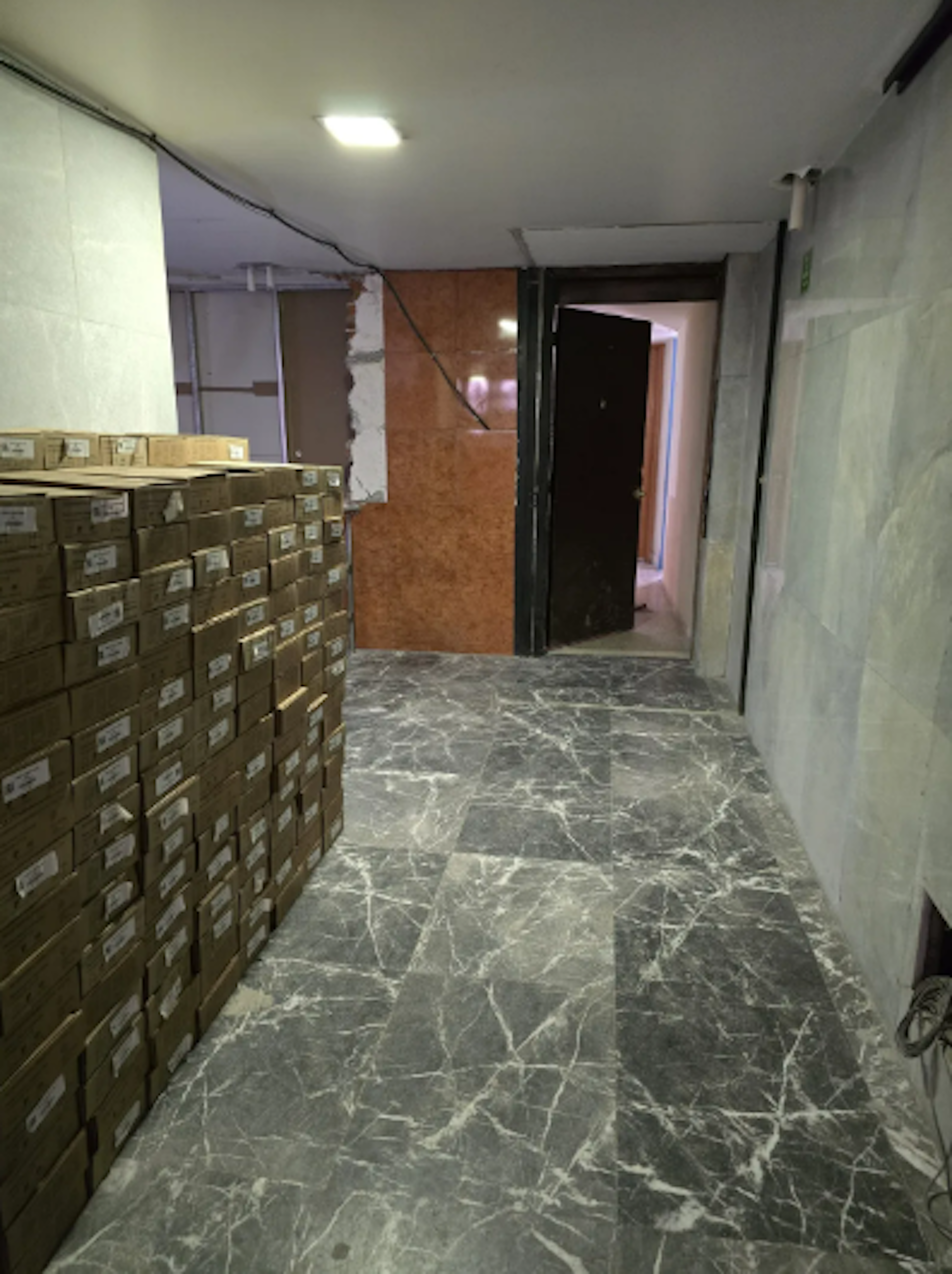 La imagen muestra un pasillo en un edificio en construcción o remodelación. El suelo es de mármol gris veteado. A la izquierda hay una pila alta de cajas de cartón etiquetadas. Las paredes están parcialmente terminadas, con algunas áreas expuestas y otras cubiertas. Se ve una puerta negra al fondo y cables eléctricos colgando del techo. El espacio parece ser un área de almacenamiento o distribución en desarrollo.