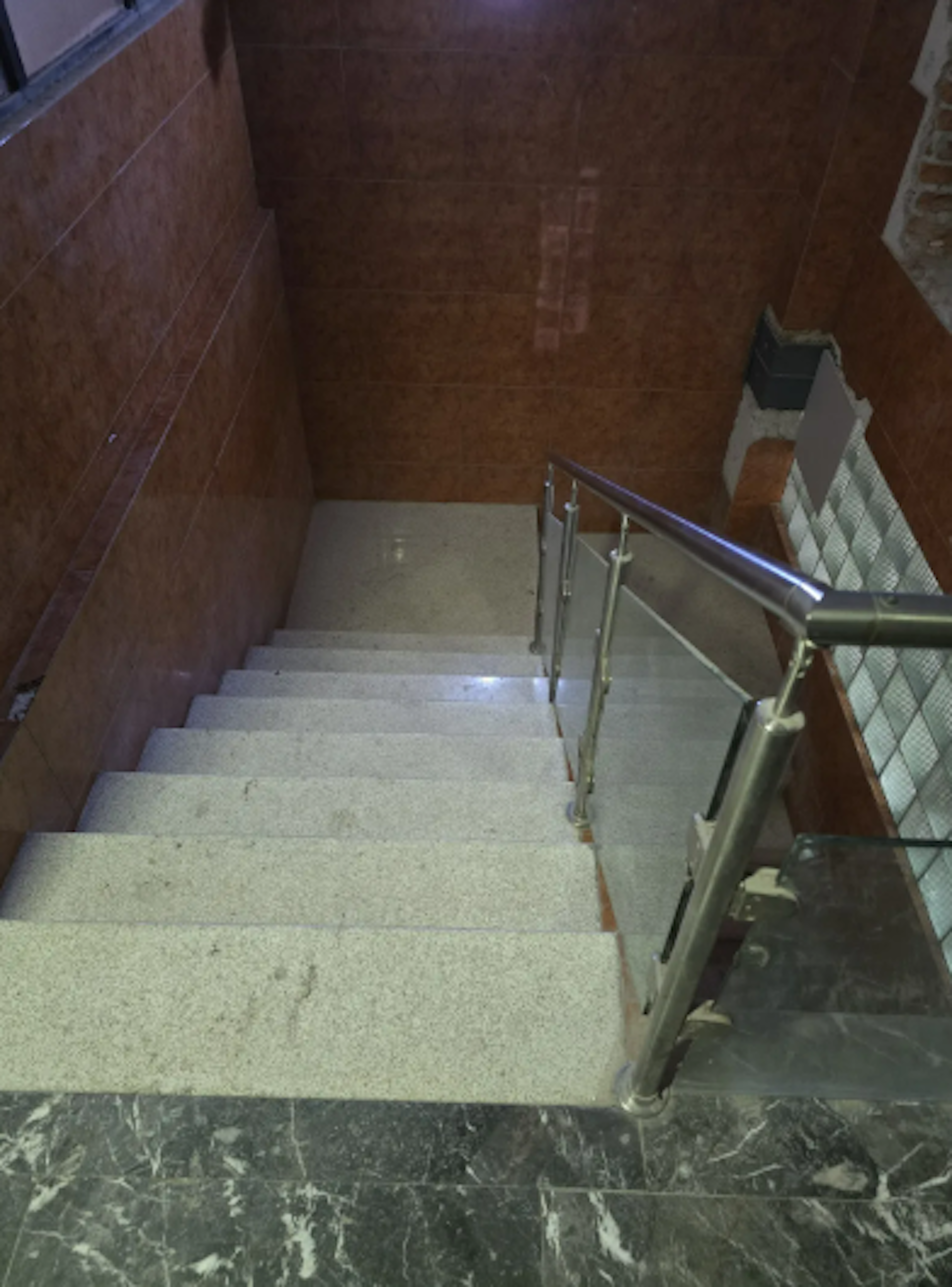 La imagen muestra una escalera interior con peldaños de mármol claro y una barandilla moderna de metal y vidrio. Las paredes están revestidas de azulejos marrones, creando un aspecto cálido y elegante. El piso inferior parece ser de mármol oscuro, contrastando con los escalones. La iluminación tenue crea un ambiente acogedor. Esta escalera refleja un diseño contemporáneo y de alta calidad en una propiedad residencial o comercial.