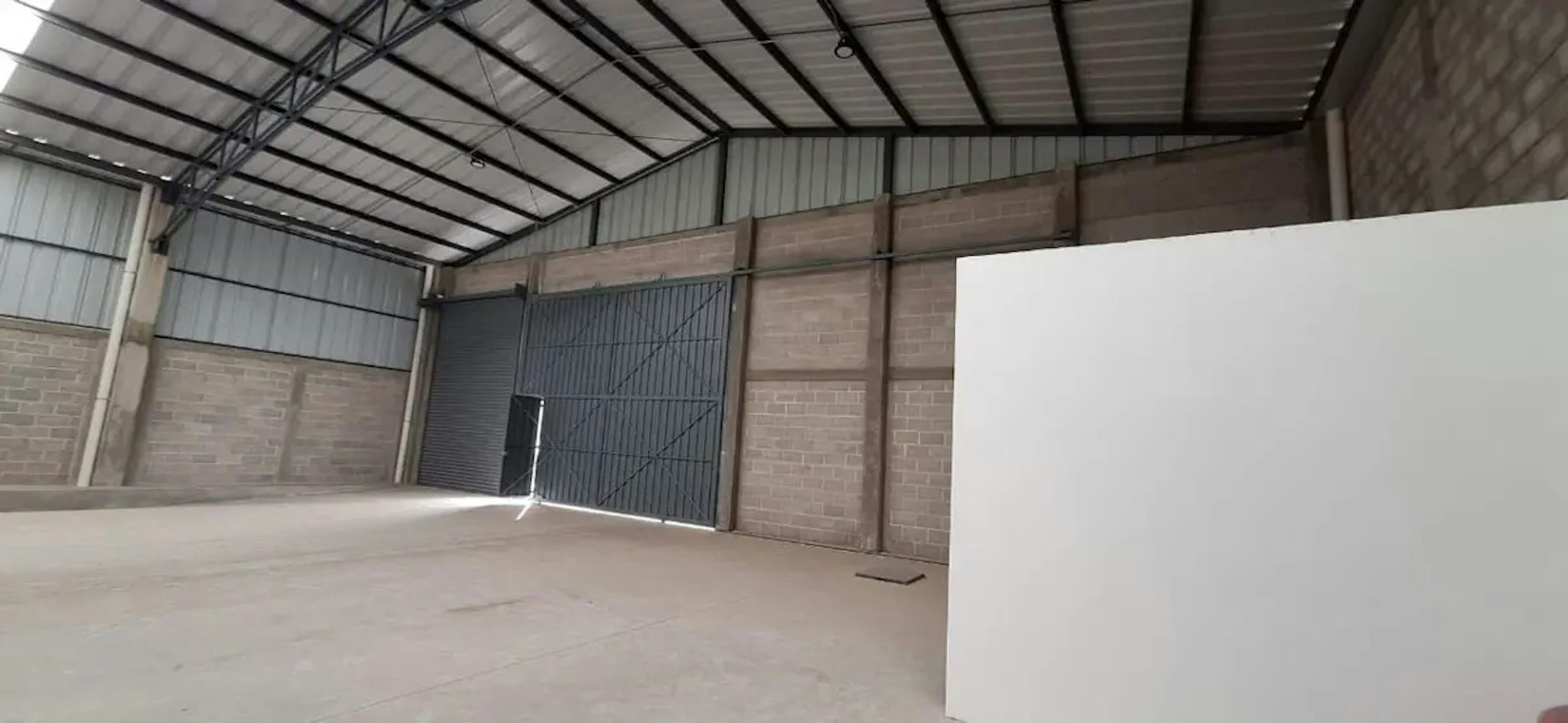 La imagen muestra el interior de una bodega o nave industrial vacía. Tiene techo alto de metal, paredes de ladrillo y concreto, y un piso de cemento limpio. Se aprecia una gran puerta metálica parcialmente abierta, permitiendo la entrada de luz natural. El espacio es amplio y diáfano, ideal para almacenamiento o uso industrial. La estructura parece estar en buenas condiciones y lista para ser ocupada.