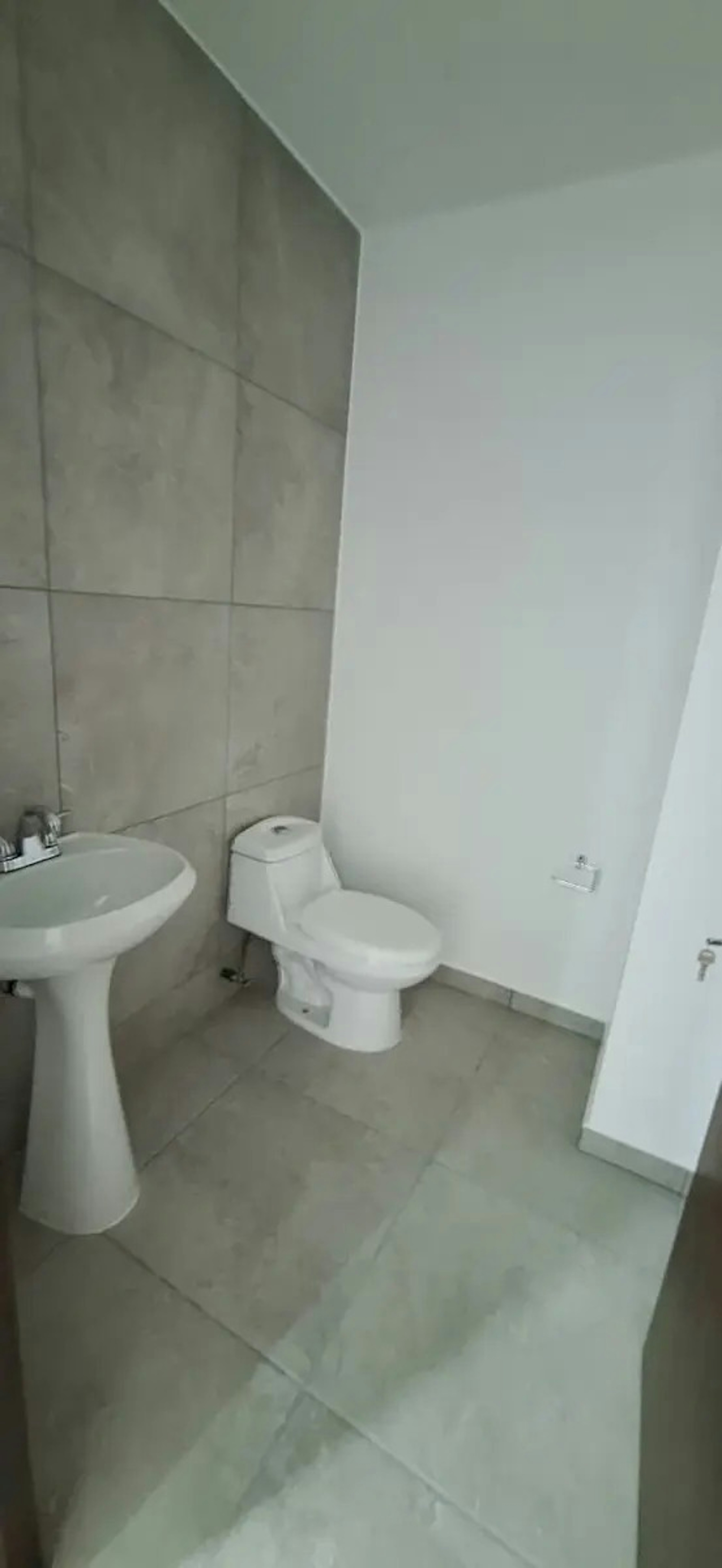 La imagen muestra un baño pequeño y moderno con paredes de azulejos grises y blancos. Cuenta con un inodoro blanco y un lavamanos de pedestal. El diseño es minimalista y funcional, típico de apartamentos o casas nuevas. El espacio parece estar recién terminado o renovado, con acabados limpios y contemporáneos. La iluminación natural es limitada, dando un aspecto fresco pero algo austero al ambiente.