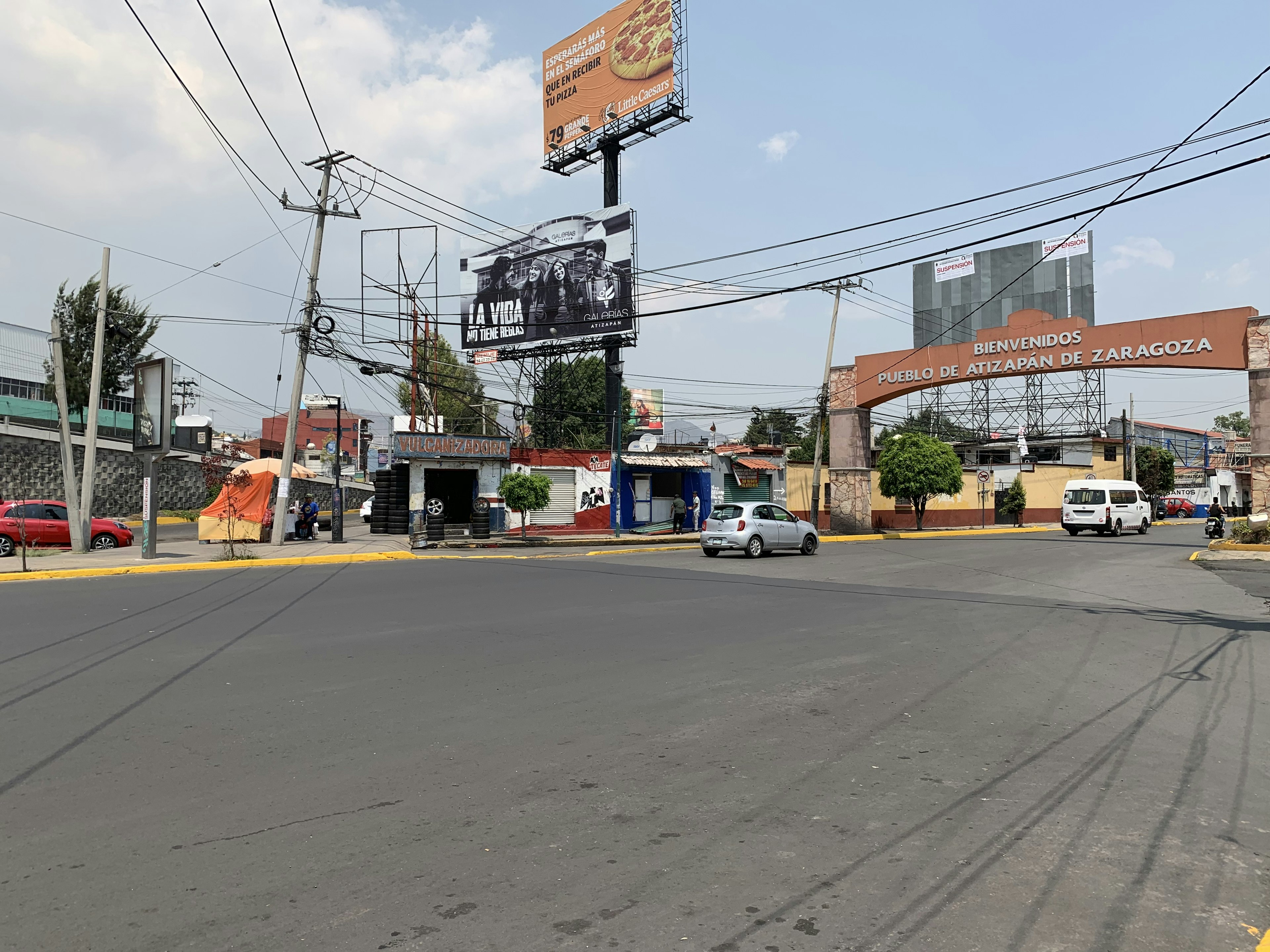 La imagen muestra una entrada a una zona urbana de México, con un letrero que dice Bienvenidos Pueblo de Atizapán de Zaragoza. Se observan edificios de baja altura, vallas publicitarias, postes de electricidad y cables. La infraestructura parece algo desgastada, con calles amplias y algunos negocios pequeños. Este entorno sugiere un área residencial y comercial de clase media-baja con potencial para desarrollo inmobiliario.