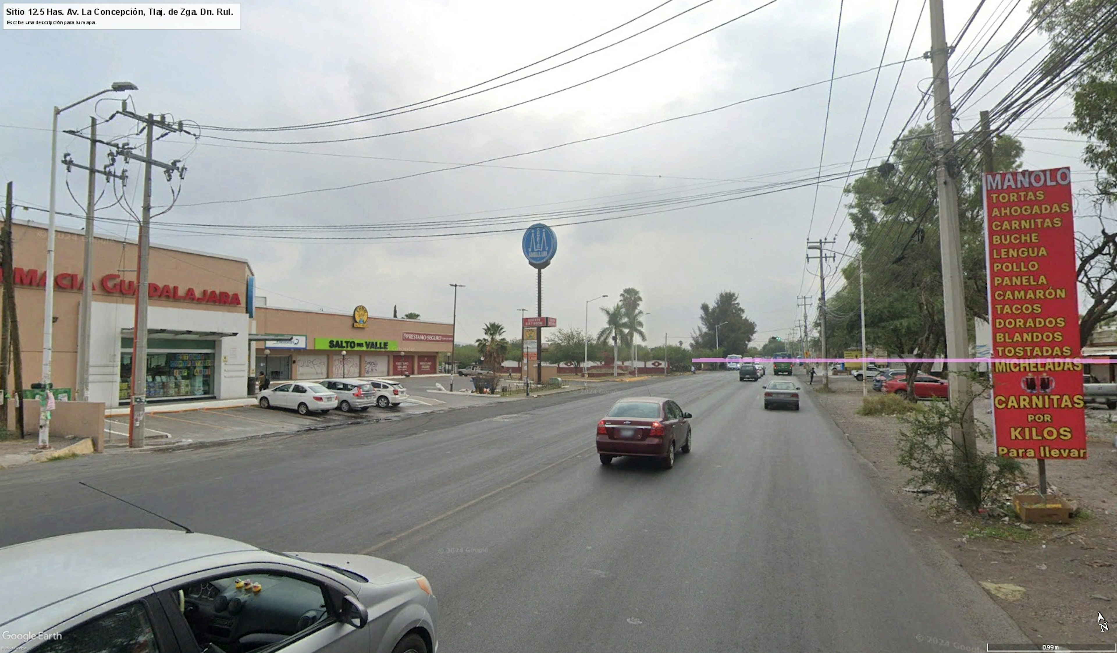 La imagen muestra una calle comercial en una zona urbana, con varios negocios y locales a lo largo de la vía. Se observan edificios de baja altura, cables eléctricos y postes de luz. Hay estacionamientos y vehículos circulando. La zona parece ser de uso mixto, combinando áreas comerciales y posiblemente residenciales. El entorno sugiere una zona en desarrollo con potencial para inversiones inmobiliarias.