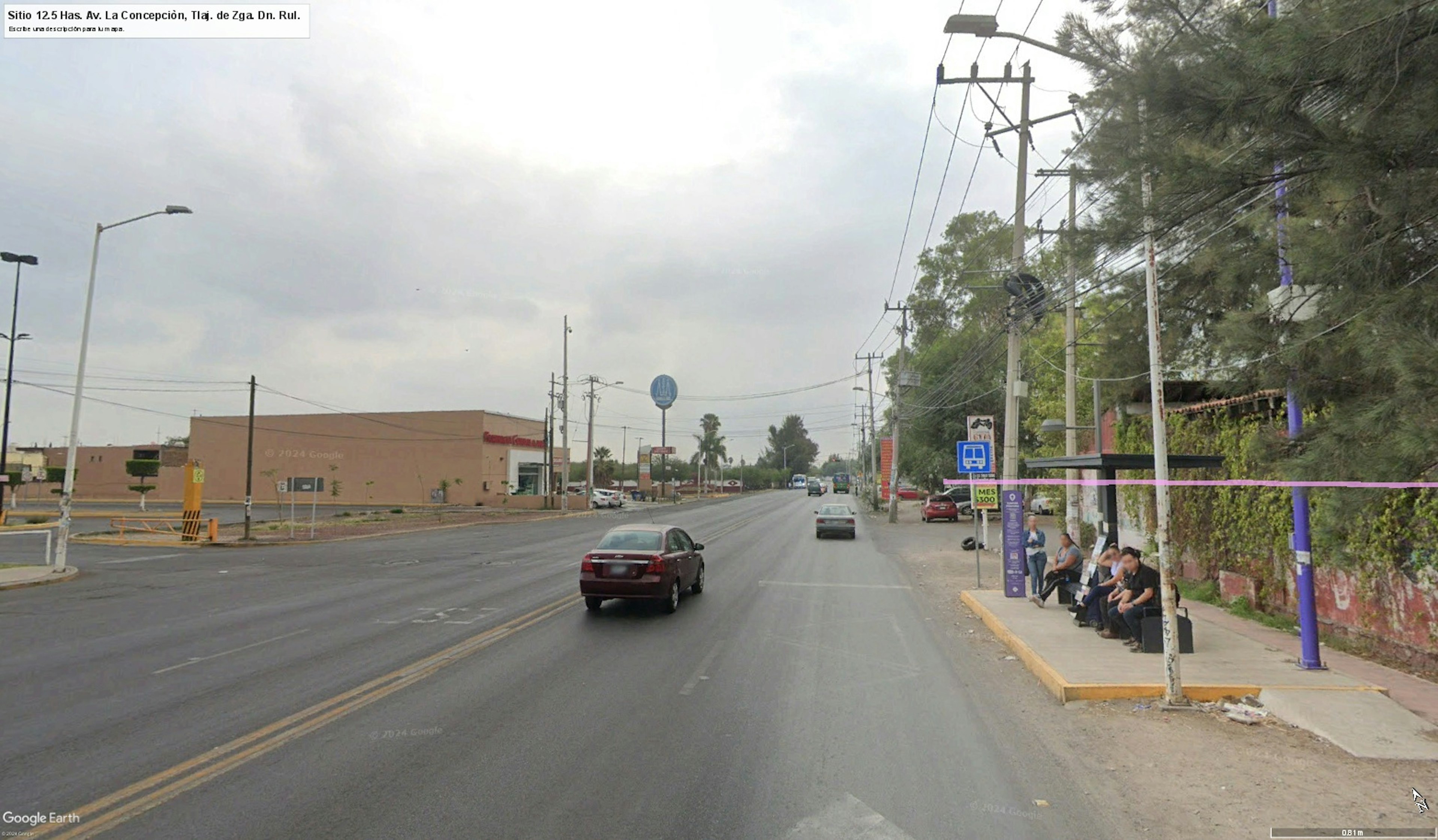 La imagen muestra una calle urbana con edificios comerciales y residenciales. Se observa una avenida de varios carriles con tráfico ligero y aceras a ambos lados. Hay postes de electricidad, señales de tráfico y una parada de autobús. El área parece estar en desarrollo, con una mezcla de construcciones nuevas y antiguas. La escena sugiere una zona en transición con potencial para futuro crecimiento inmobiliario.