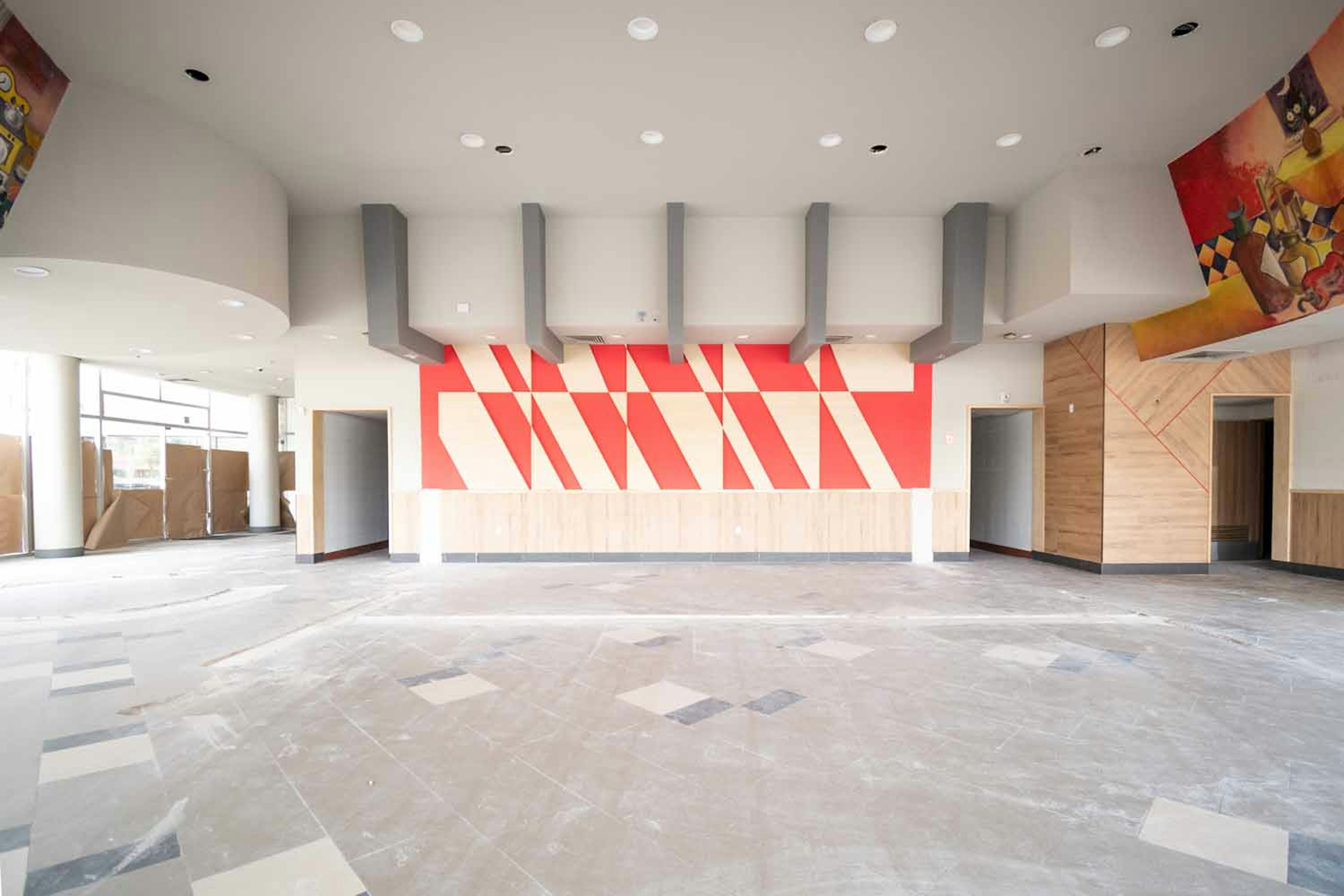 The image shows a modern, spacious interior of a building, likely a commercial or institutional space. It features a striking red geometric design on the central wall, surrounded by gray pillars. The ceiling has recessed lighting, and there are artistic elements visible on the side walls. The floor appears to be in a state of construction or renovation, with a rough concrete finish. The overall design is sleek and contemporary, with clean lines and a minimalist aesthetic.