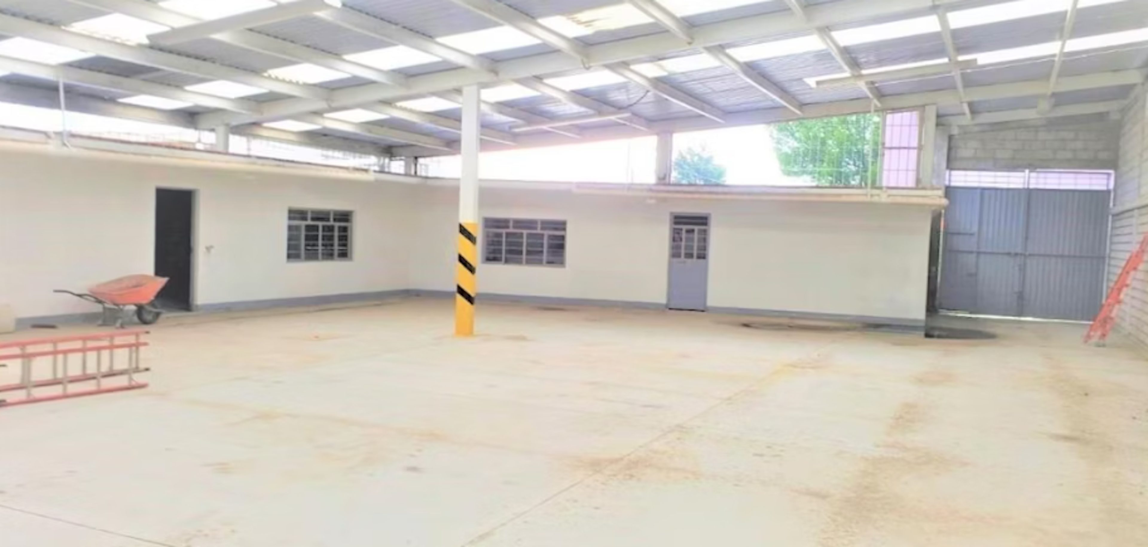 La imagen muestra un amplio espacio cubierto con techo transparente, ideal para un garaje o área de almacenamiento. Tiene paredes blancas, piso de concreto y varias puertas y ventanas. Se observa una carretilla naranja y una escalera, sugiriendo un uso industrial o de taller. El espacio es luminoso y ofrece potencial para diversos usos comerciales o de almacenamiento.