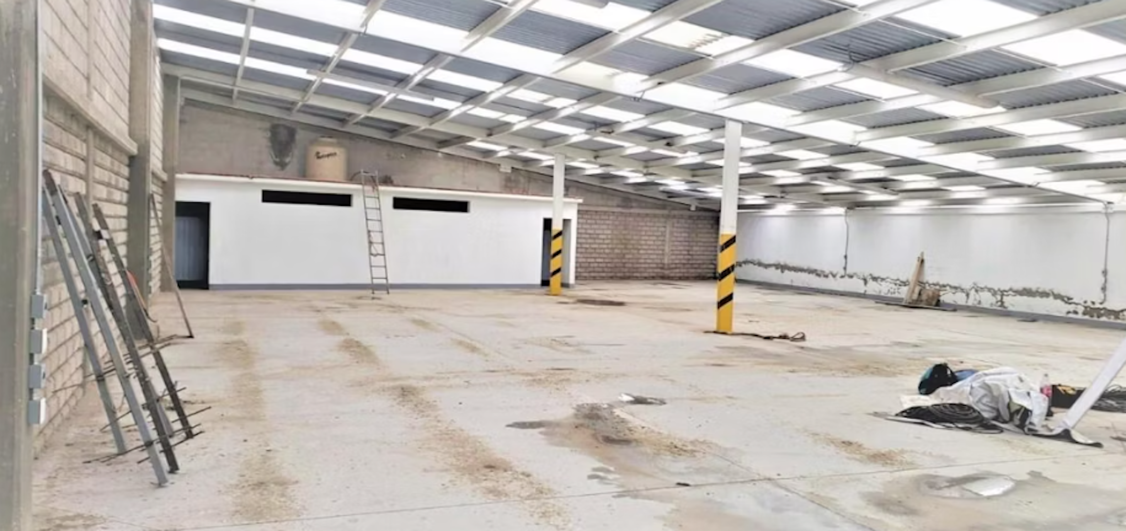 La imagen muestra un amplio espacio industrial en proceso de renovación. Se observa un techo metálico con iluminación, paredes de ladrillo y blancas, y un piso de concreto. Hay escaleras, postes de seguridad amarillos y negros, y algunos materiales de construcción dispersos. El área parece estar siendo preparada para un nuevo uso, posiblemente como almacén, taller o espacio comercial.